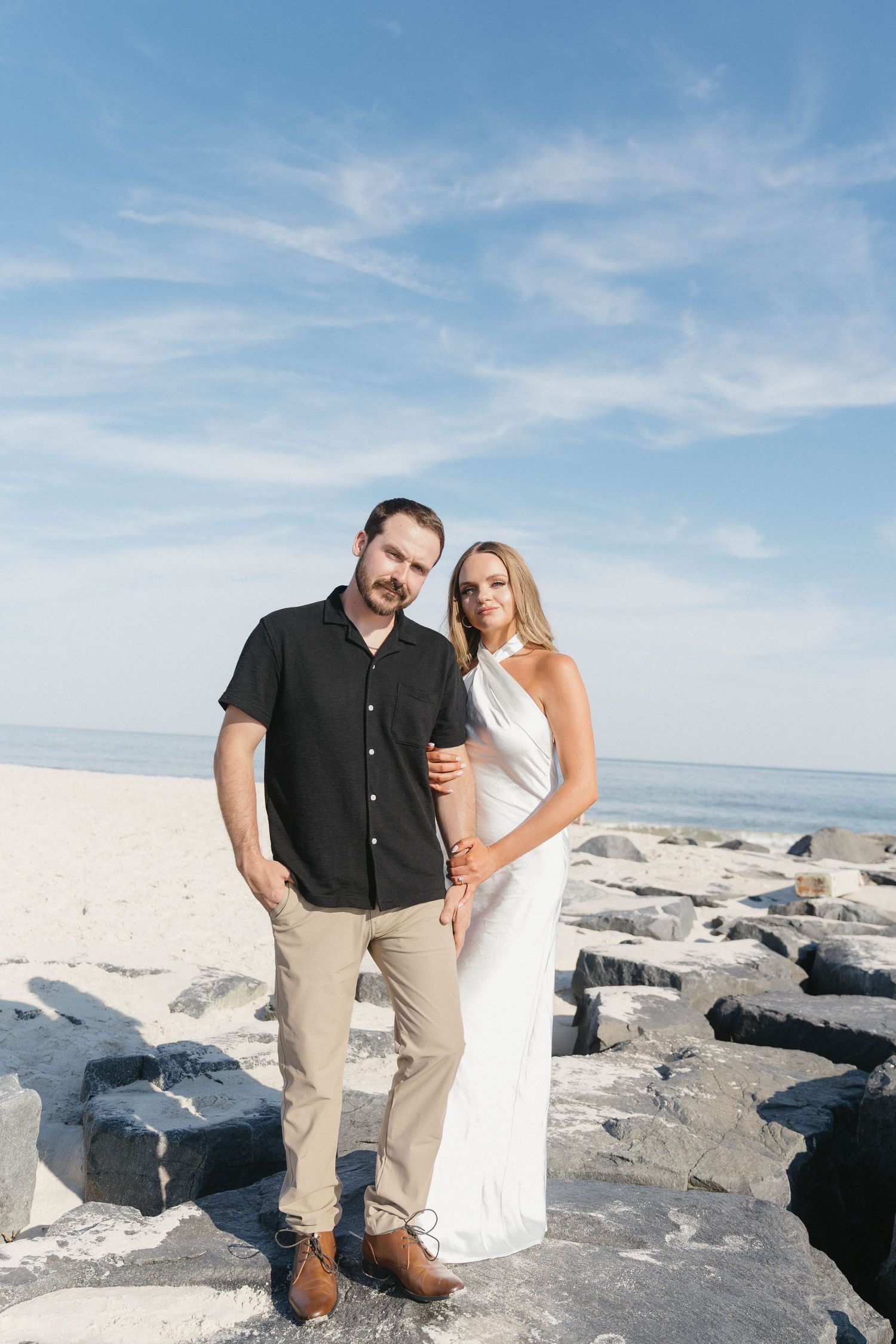 A couple poses on rocky beach, man in black shirt and khakis, woman in white halter dress, ocean and blue sky behind them.