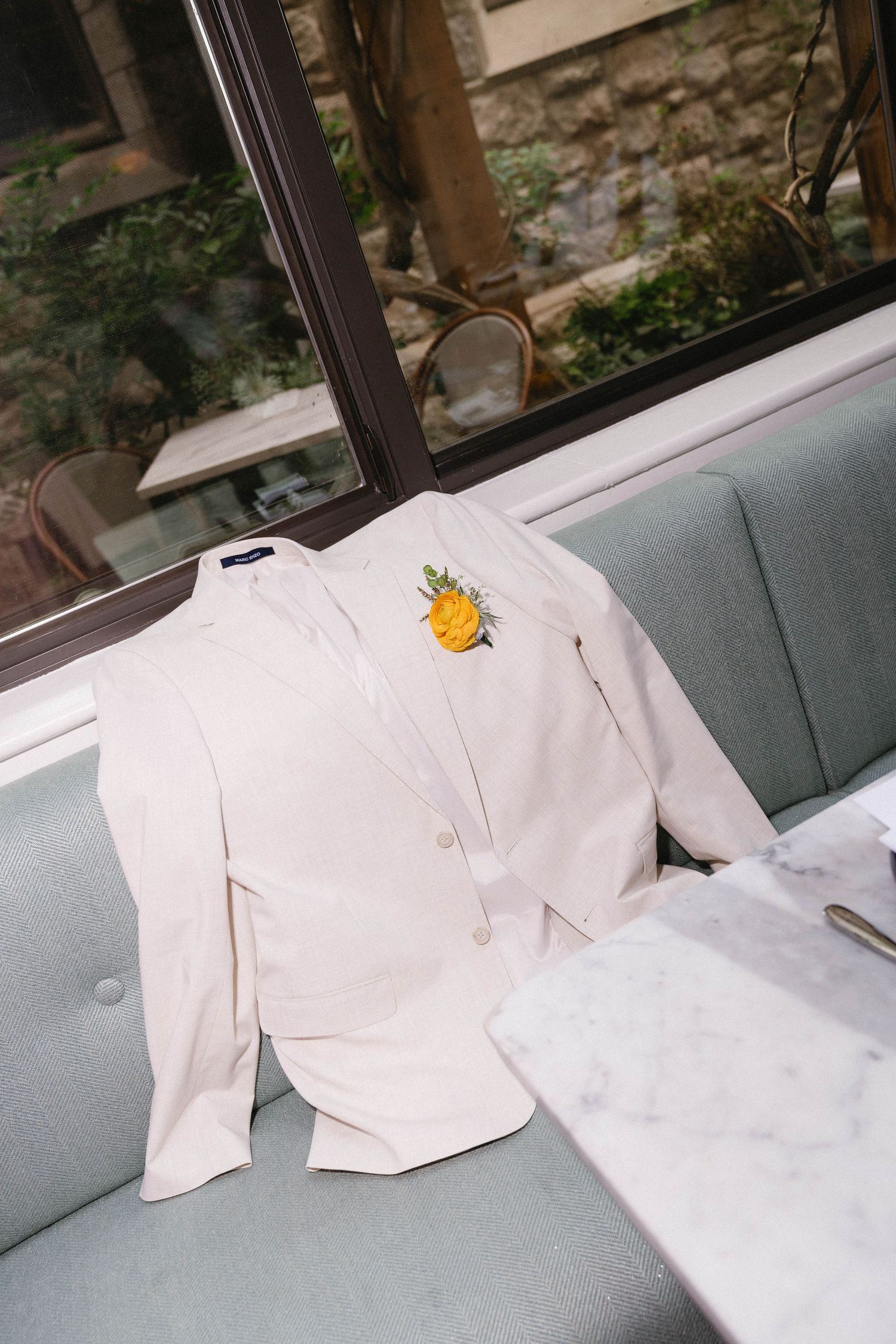 White wedding suit jacket with yellow floral boutonniere displayed on a light blue velvet restaurant booth seat.