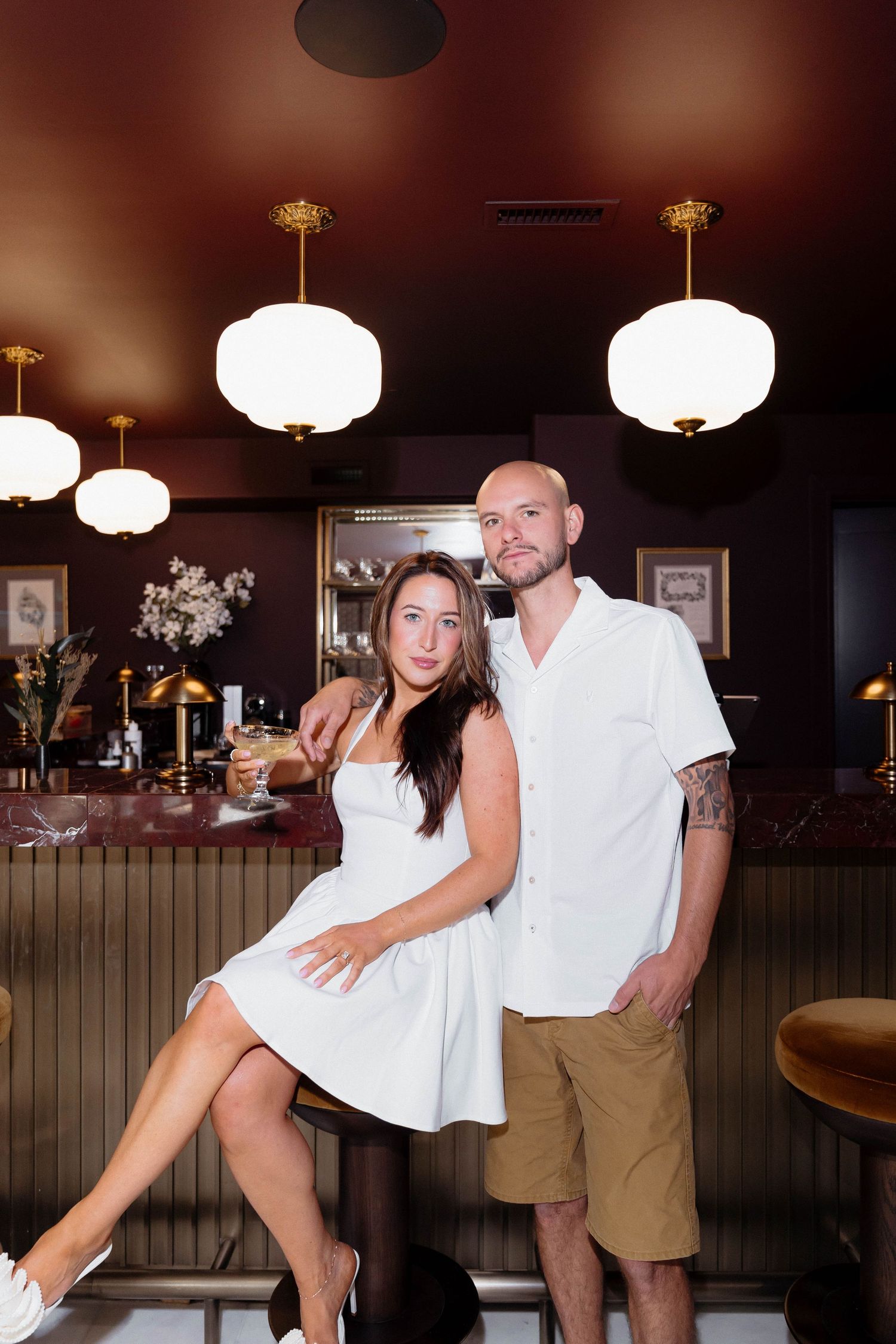 A couple in white outfits poses at an upscale bar with warm lighting and dark red decor.