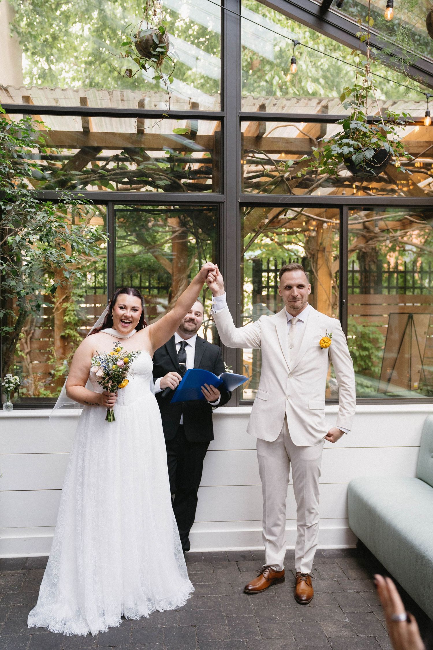 Bride and groom raise joined hands in celebration at indoor wedding ceremony with greenery backdrop.