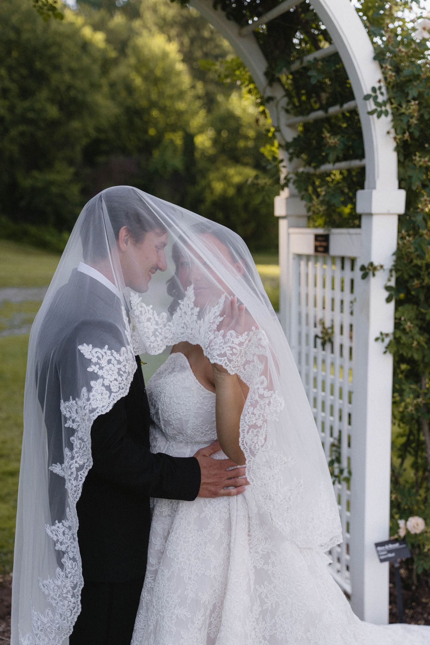 Bride and groom share romantic moment under lace veil by white garden arch surrounded by lush greenery.