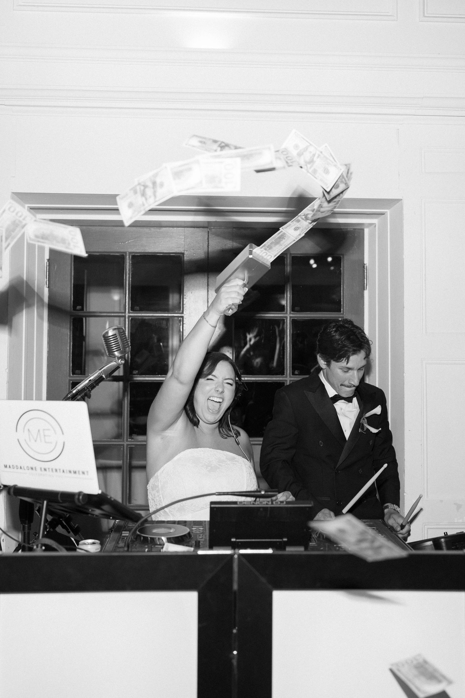 Black and white photo of a bride raising money at a wedding DJ booth beside a groom in a tuxedo.