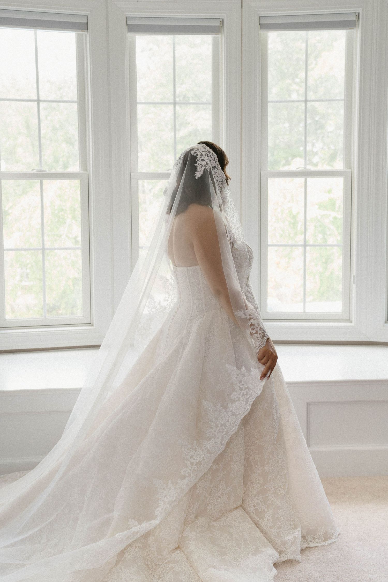 Elegant bride in lace ball gown with cathedral veil standing before bright white windows.