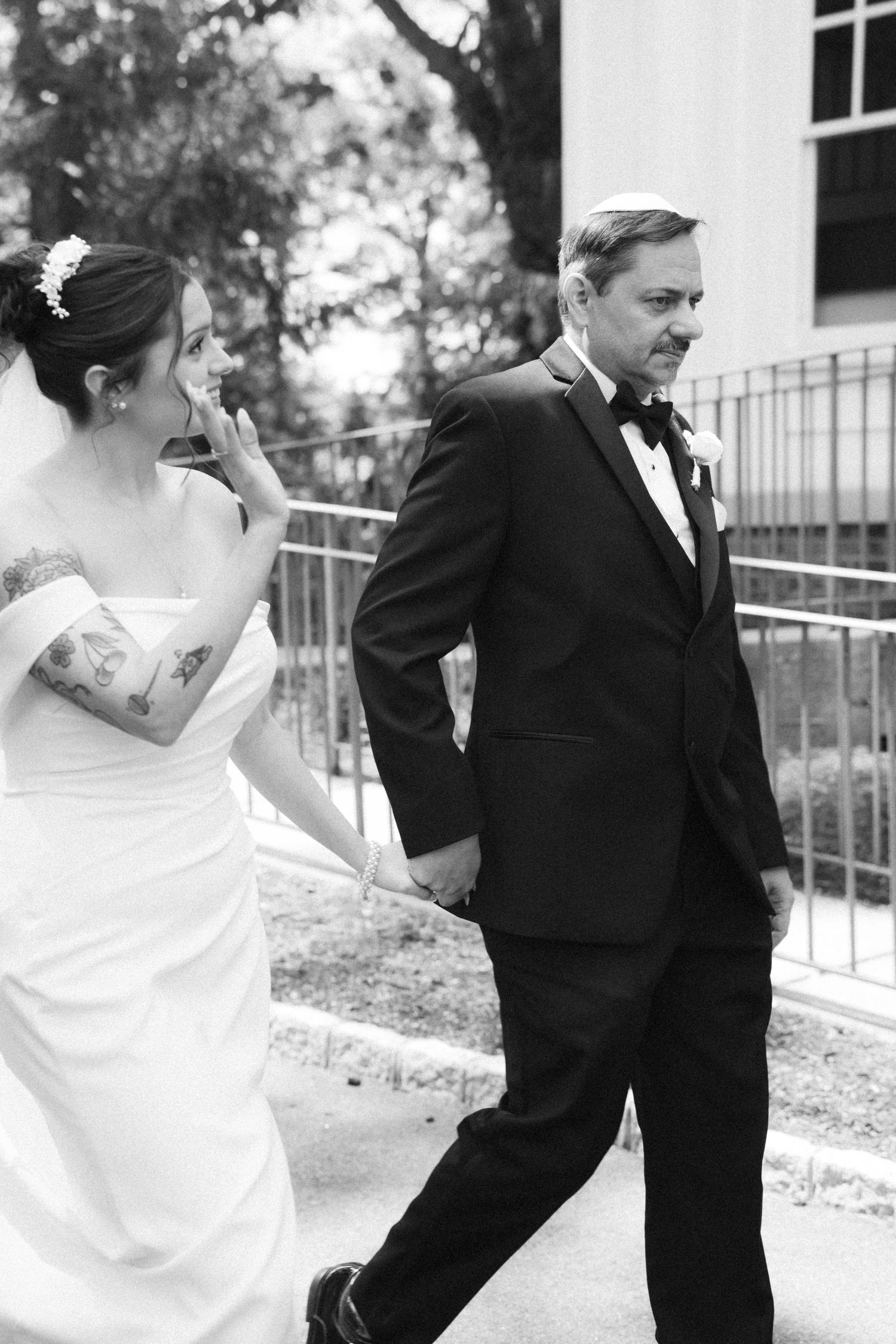 Black and white photo of tattooed bride in white gown holding hands with groom in tuxedo.