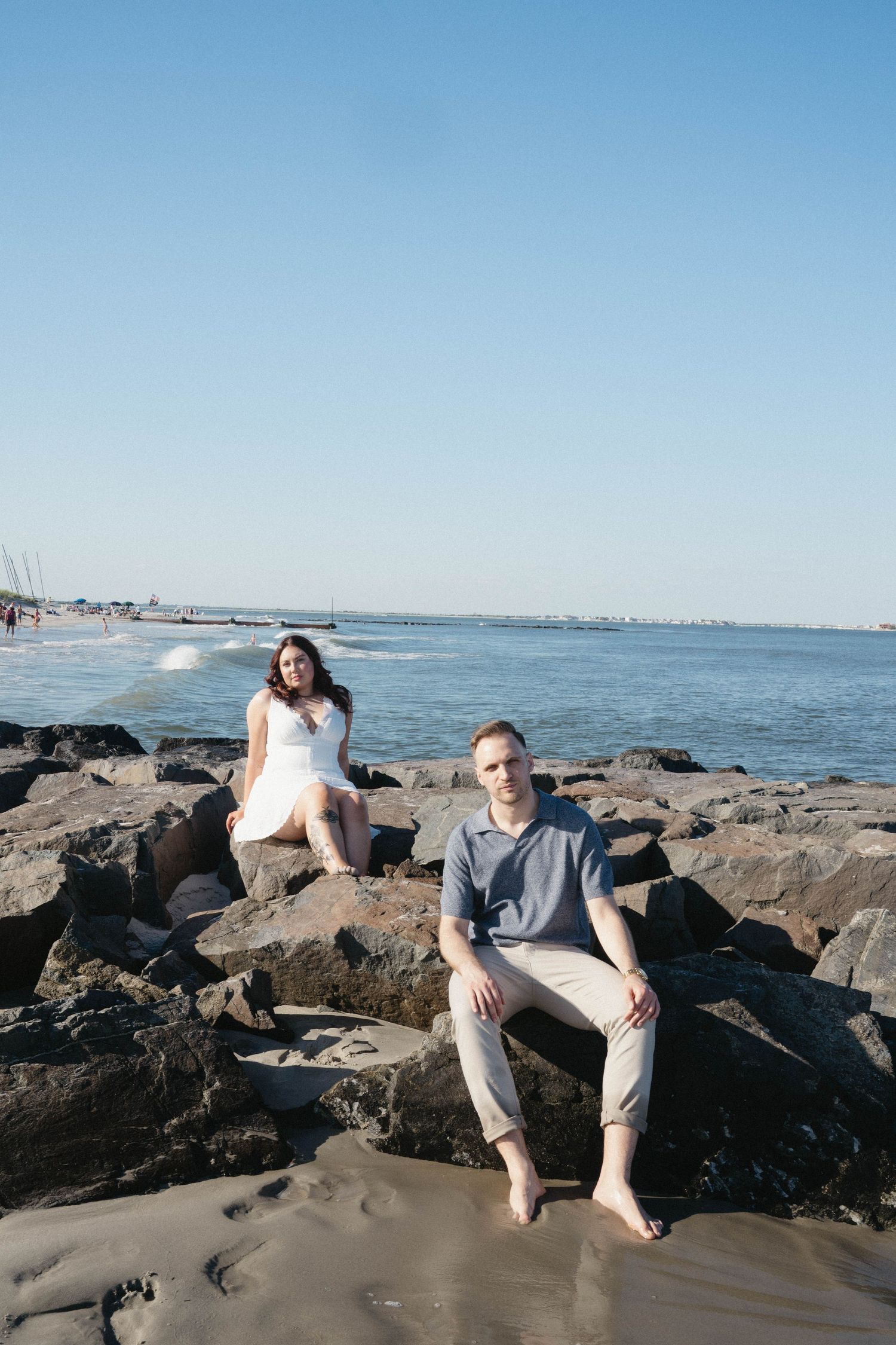 A couple sits on rocky jetty by the ocean, woman in white dress and man in blue shirt, with calm sea in background.