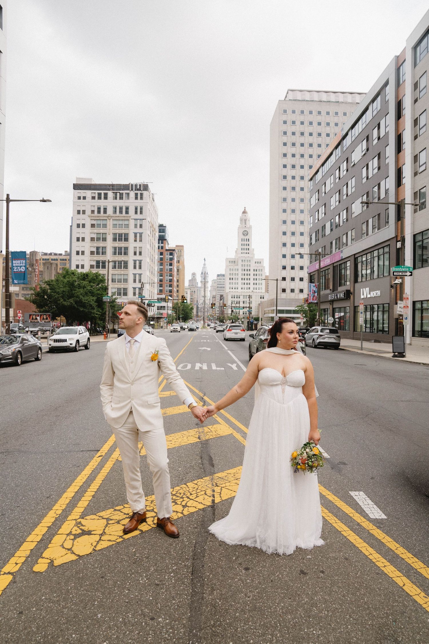 A bride and groom hold hands while crossing a city street, with urban buildings and a cloudy sky in the background.