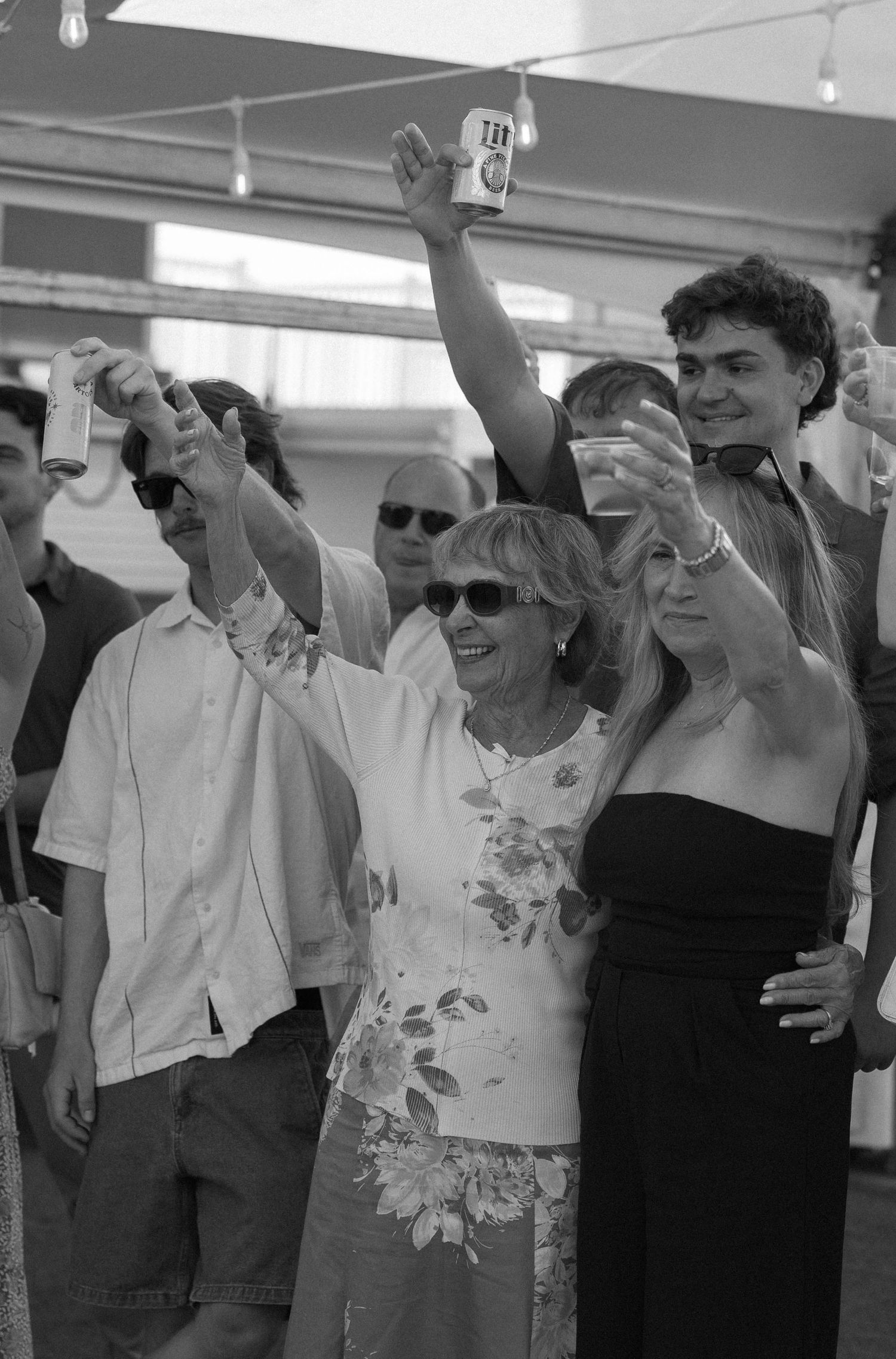 Black and white photo of guests raising drinks in a toast at a lively outdoor celebration.