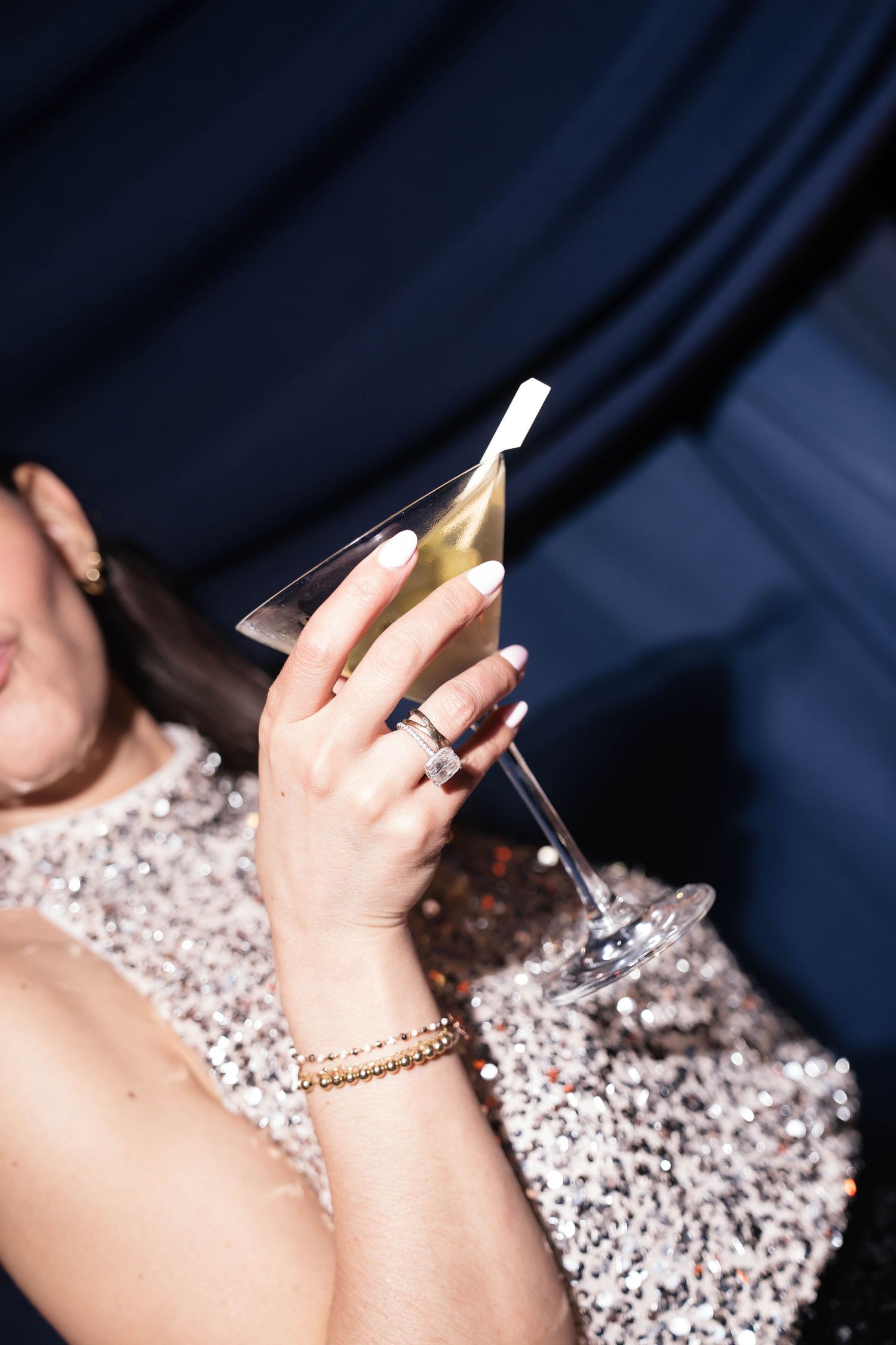 Close-up of a woman in a sequined dress holding a martini glass with diamond rings and gold bracelet.
