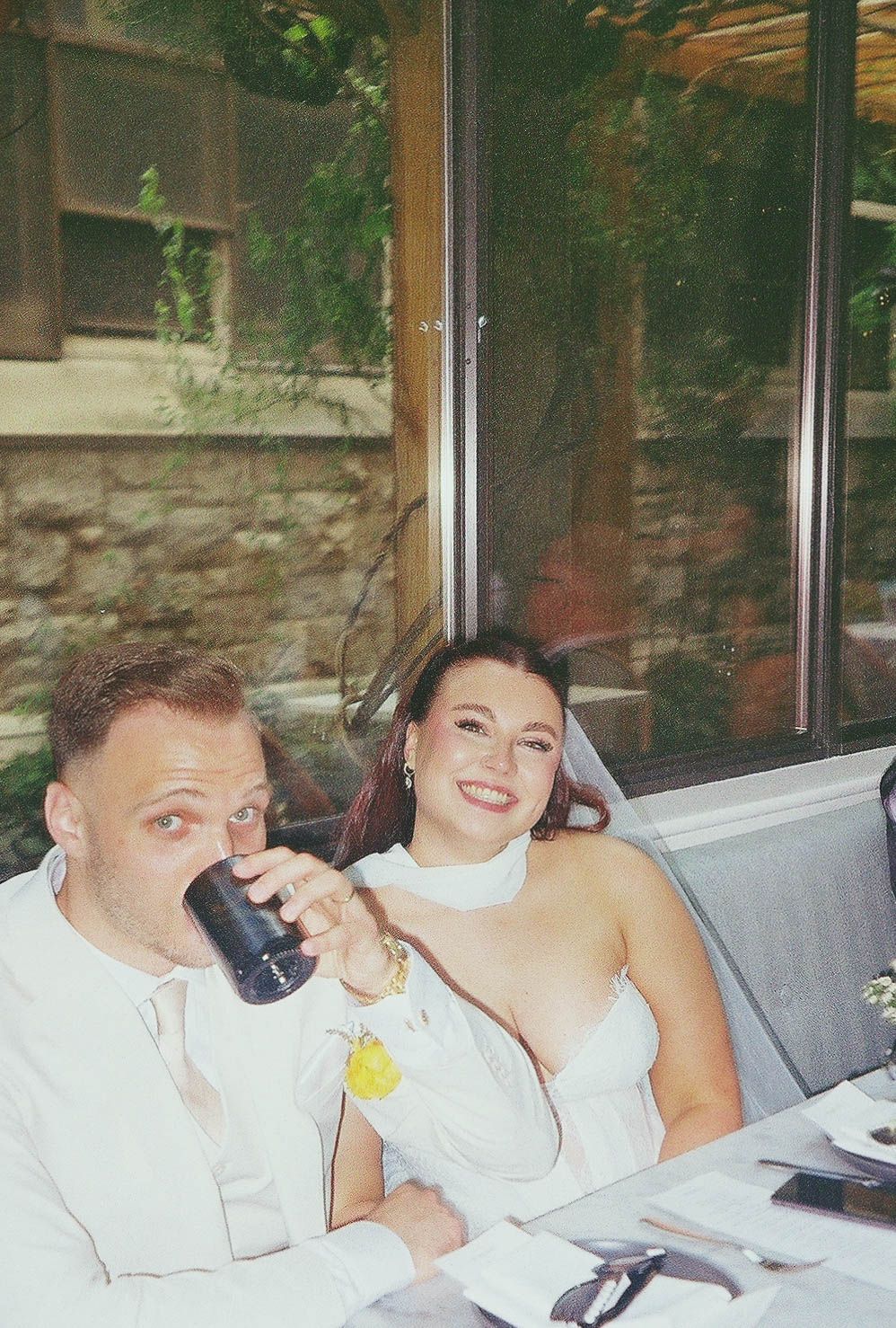 A couple in white wedding attire smiles and drinks at an outdoor reception venue with stone walls.