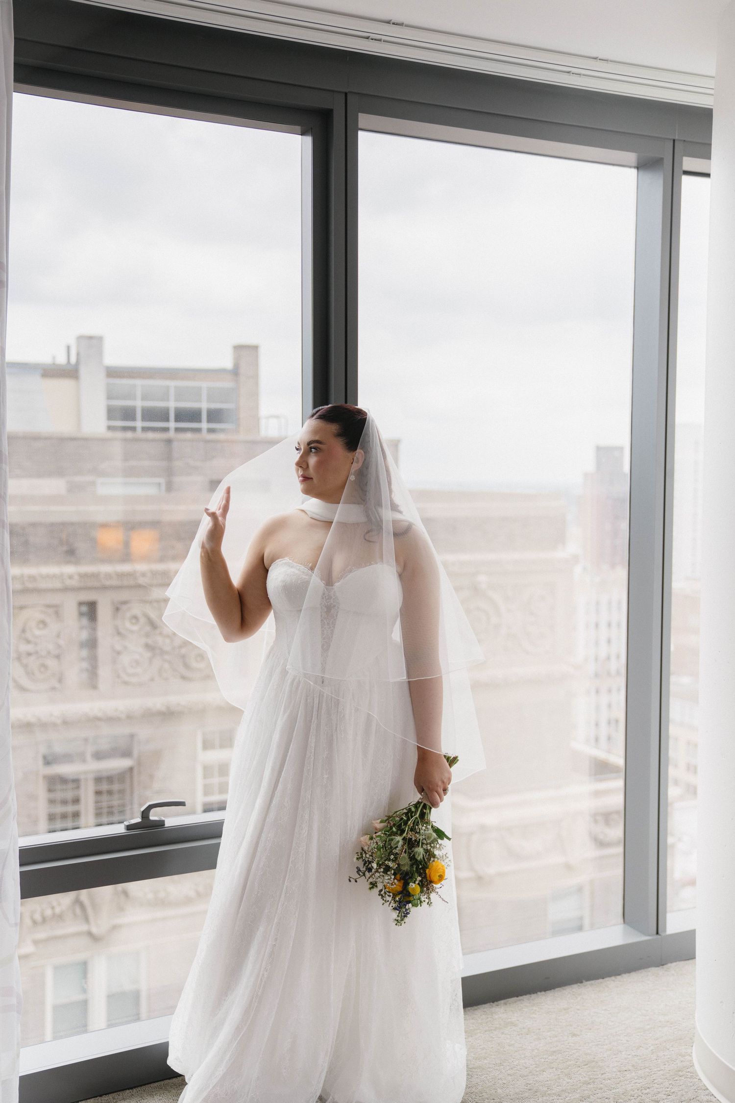 Bride in white gown and veil holds yellow bouquet by large windows with city skyline view.