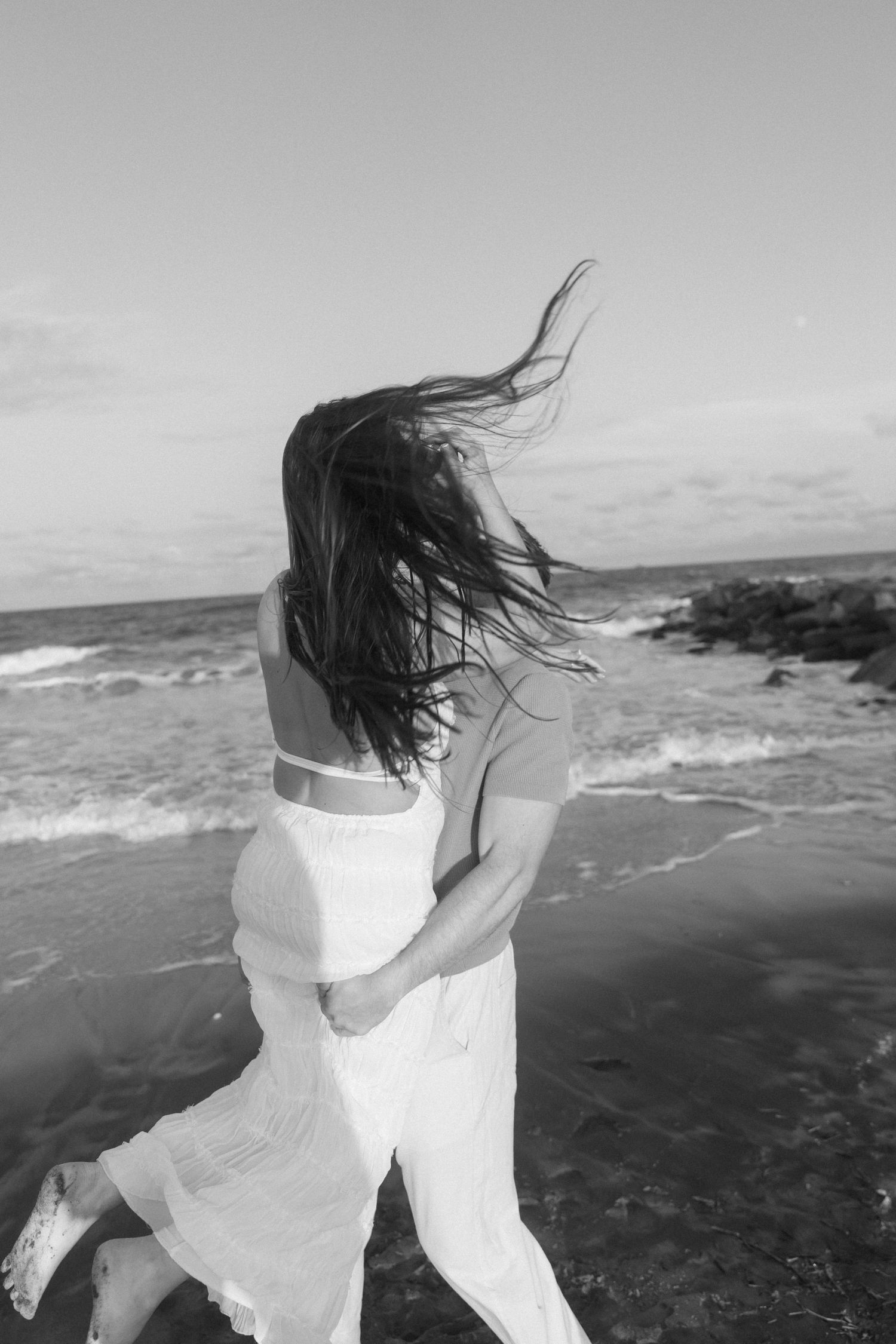 Black and white photo of a couple embracing on a windy beach with ocean waves in the background.