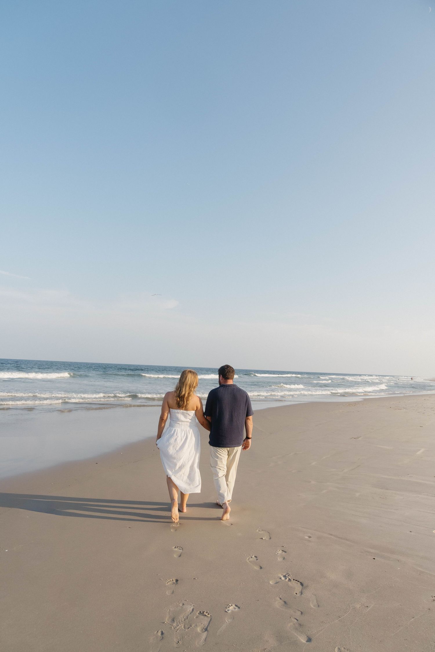 Couple walking barefoot on a sandy beach in white and navy outfits, leaving footprints with ocean waves in the background.