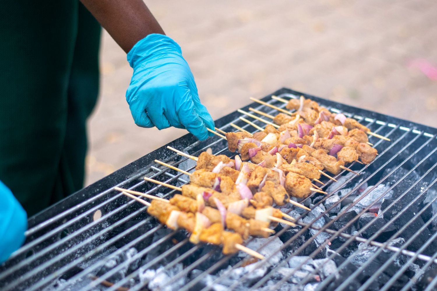 Person in blue gloves grilling seasoned meat skewers on a charcoal barbecue grill.