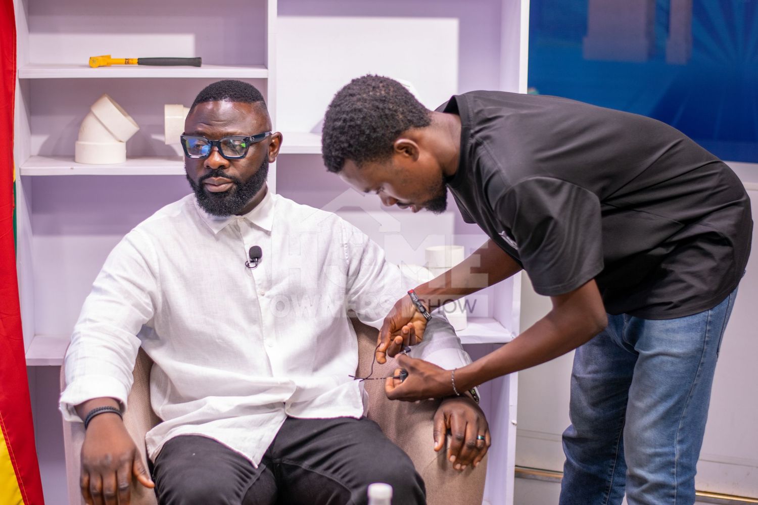 A man in a white shirt sits while another person attaches a microphone to his lapel in a TV studio.