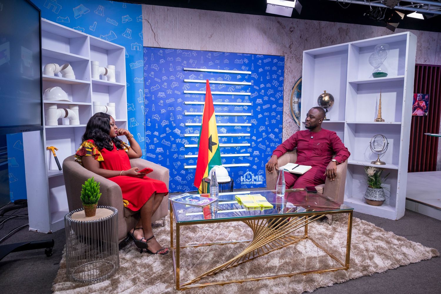 Two professionals in colorful attire sit for a TV interview in a modern studio with Ghana flag backdrop.