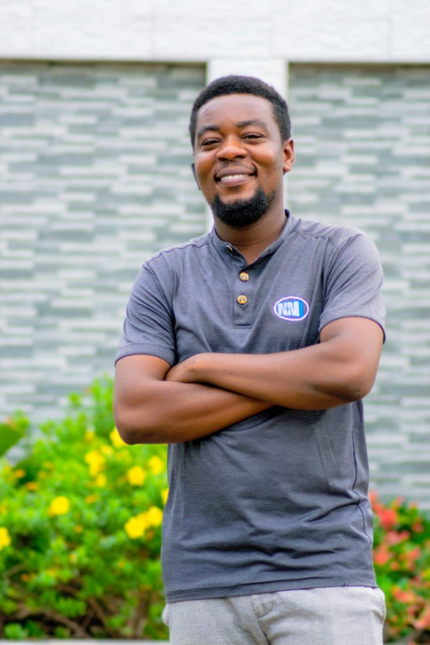Smiling African man with crossed arms wearing a gray polo shirt with logo, standing outdoors near colorful flowers.