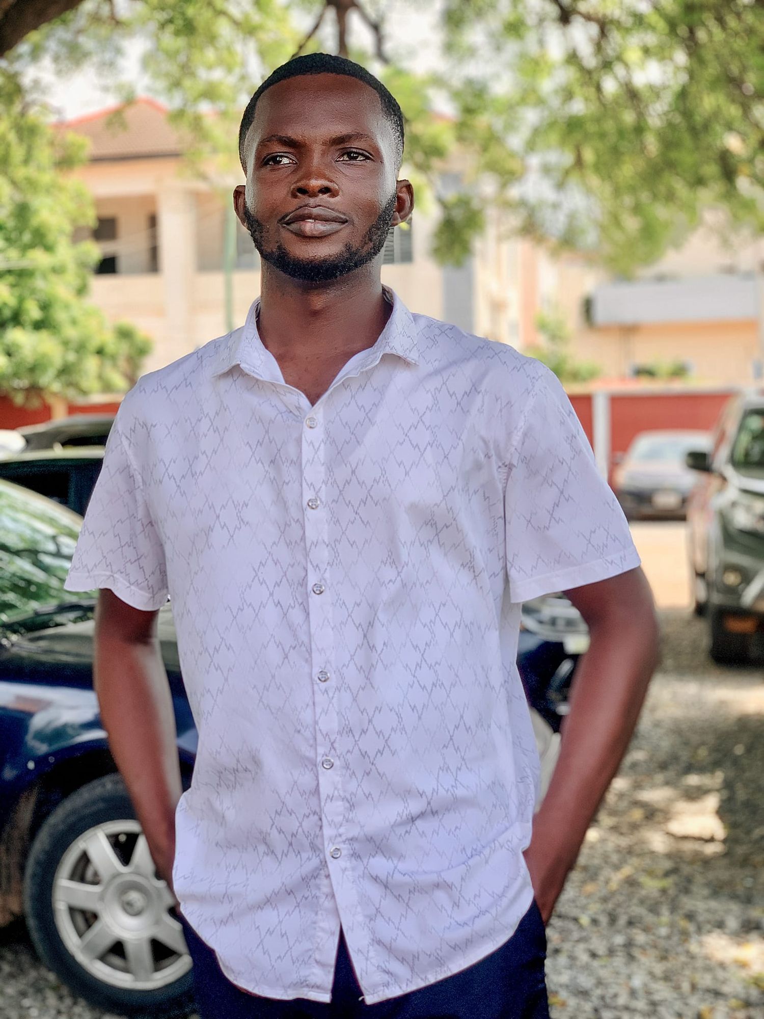 Young man in white patterned short-sleeve shirt standing outdoors near parked cars and green trees.