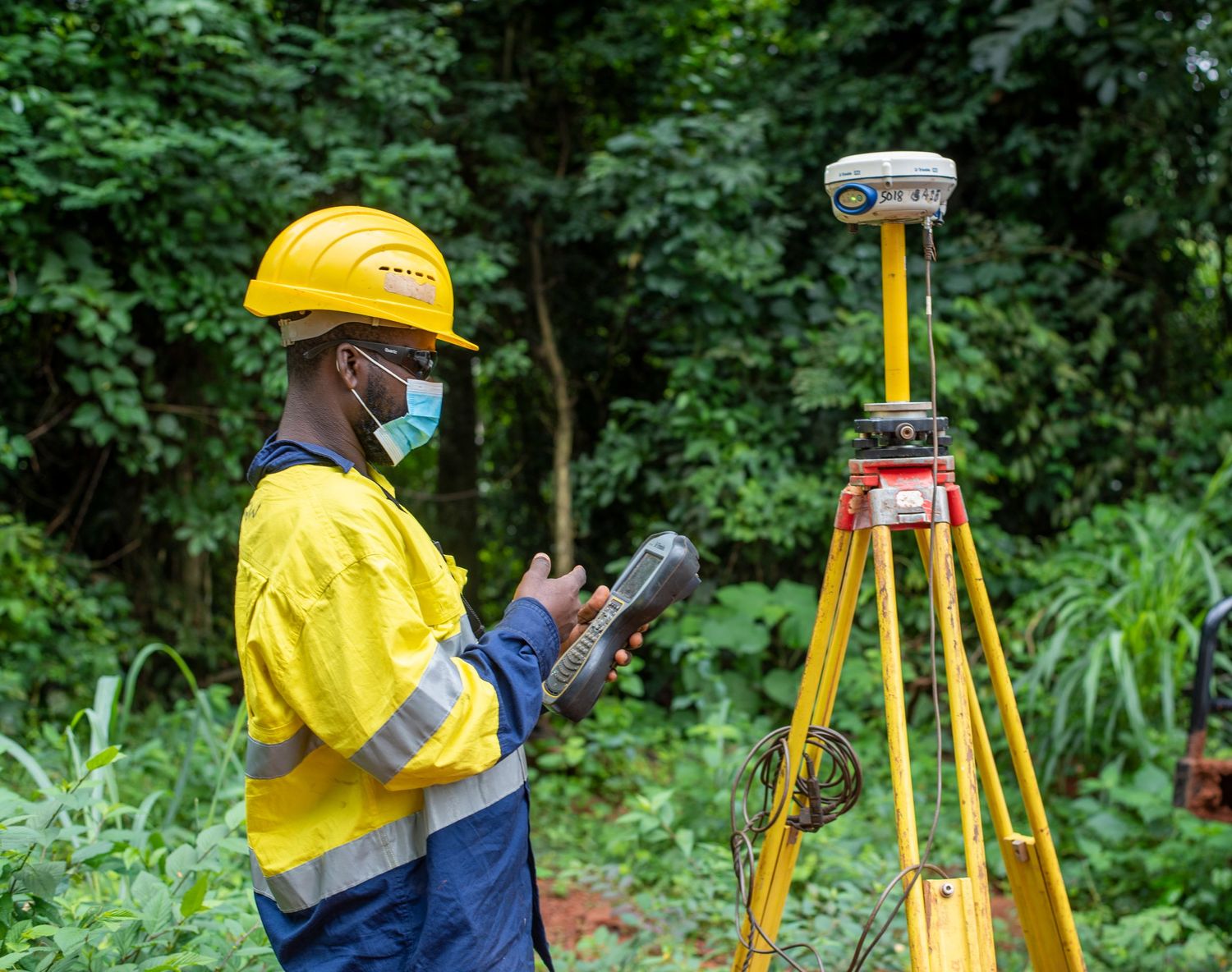 African engineer in safety gear using surveying equipment in lush green forest.