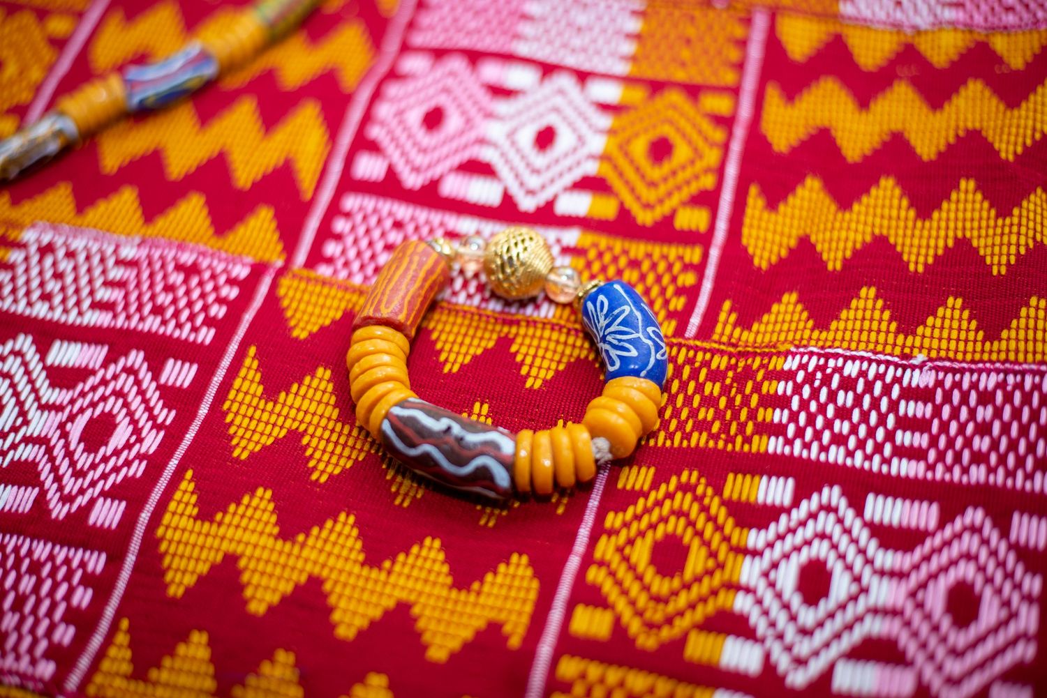 Colorful African Kente fabric in red, yellow, and white patterns with traditional beaded bracelet and pendant.