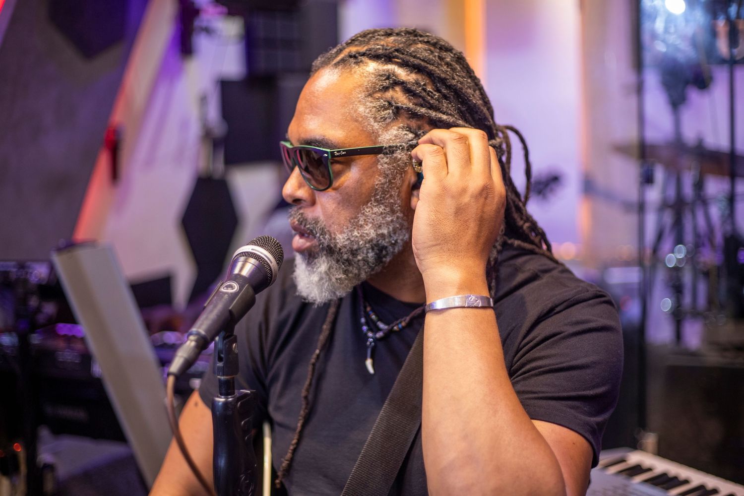 A man with dreadlocks and sunglasses adjusts headphones at a microphone in a music studio setting.
