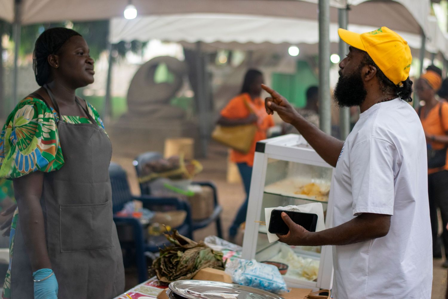 Vendor and customer conversing at an outdoor African food market stall with fresh produce.