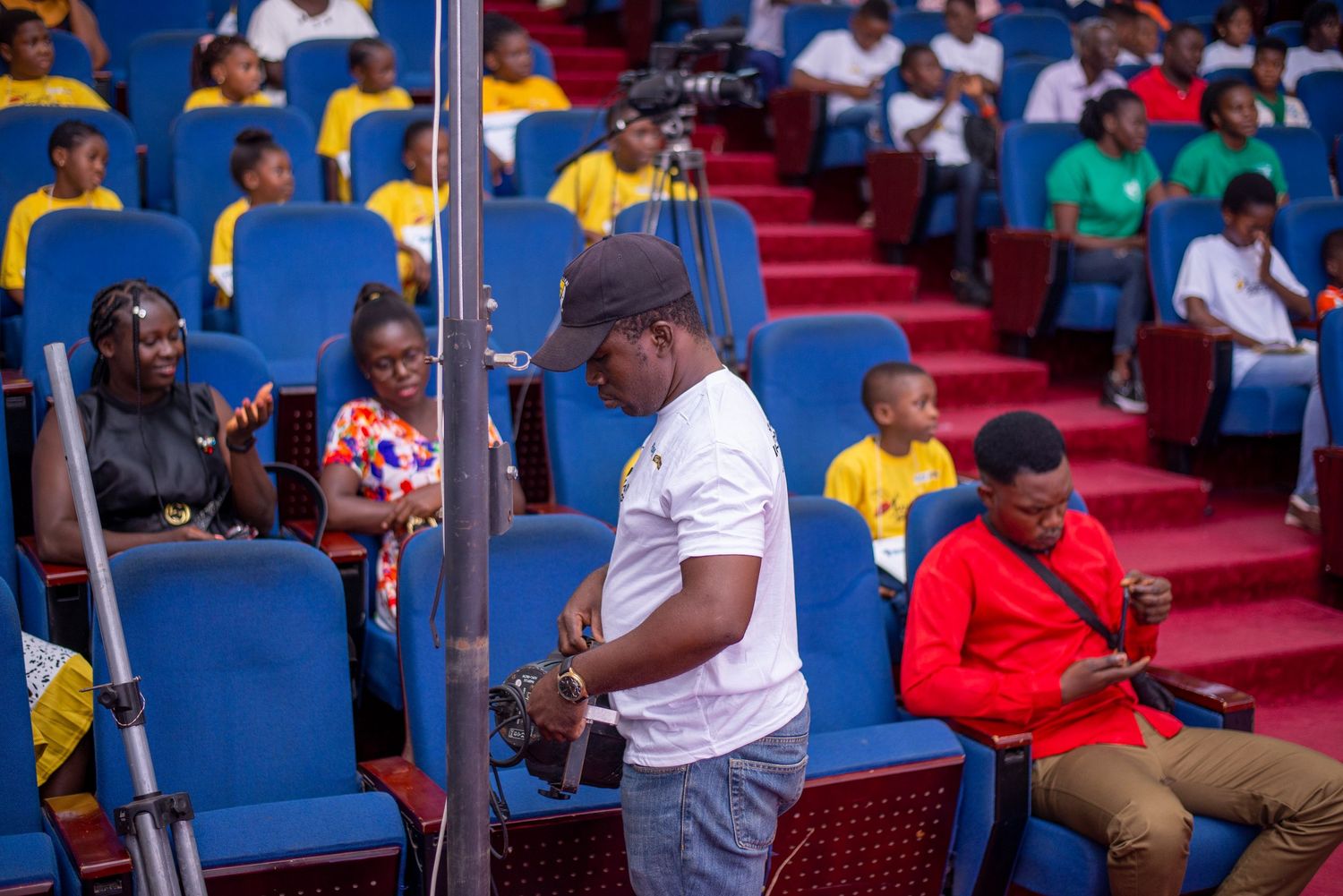 Cameraman with equipment in a colorful auditorium filled with seated audience members.