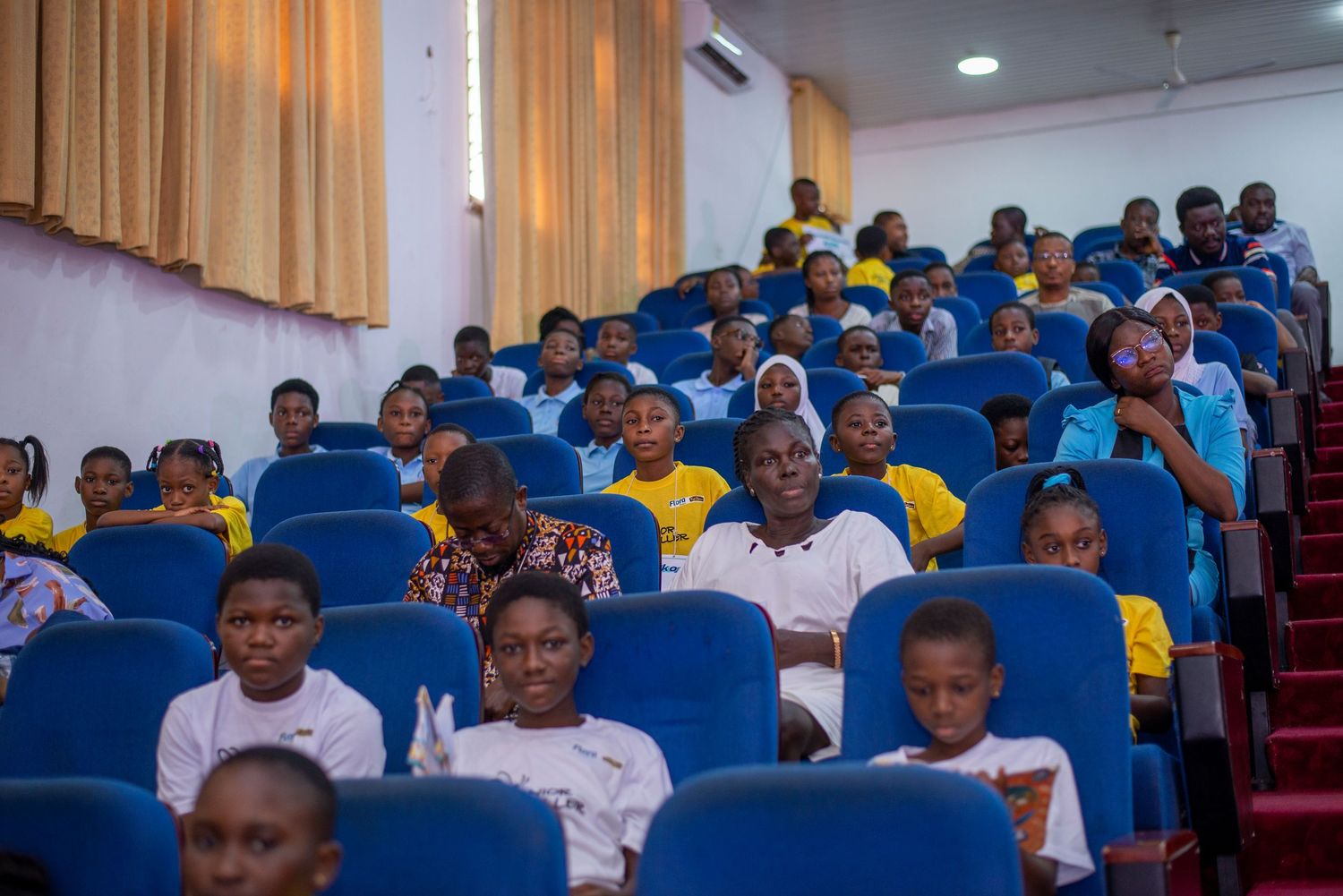 Children and adults seated in blue auditorium chairs wearing yellow and white shirts at an event.