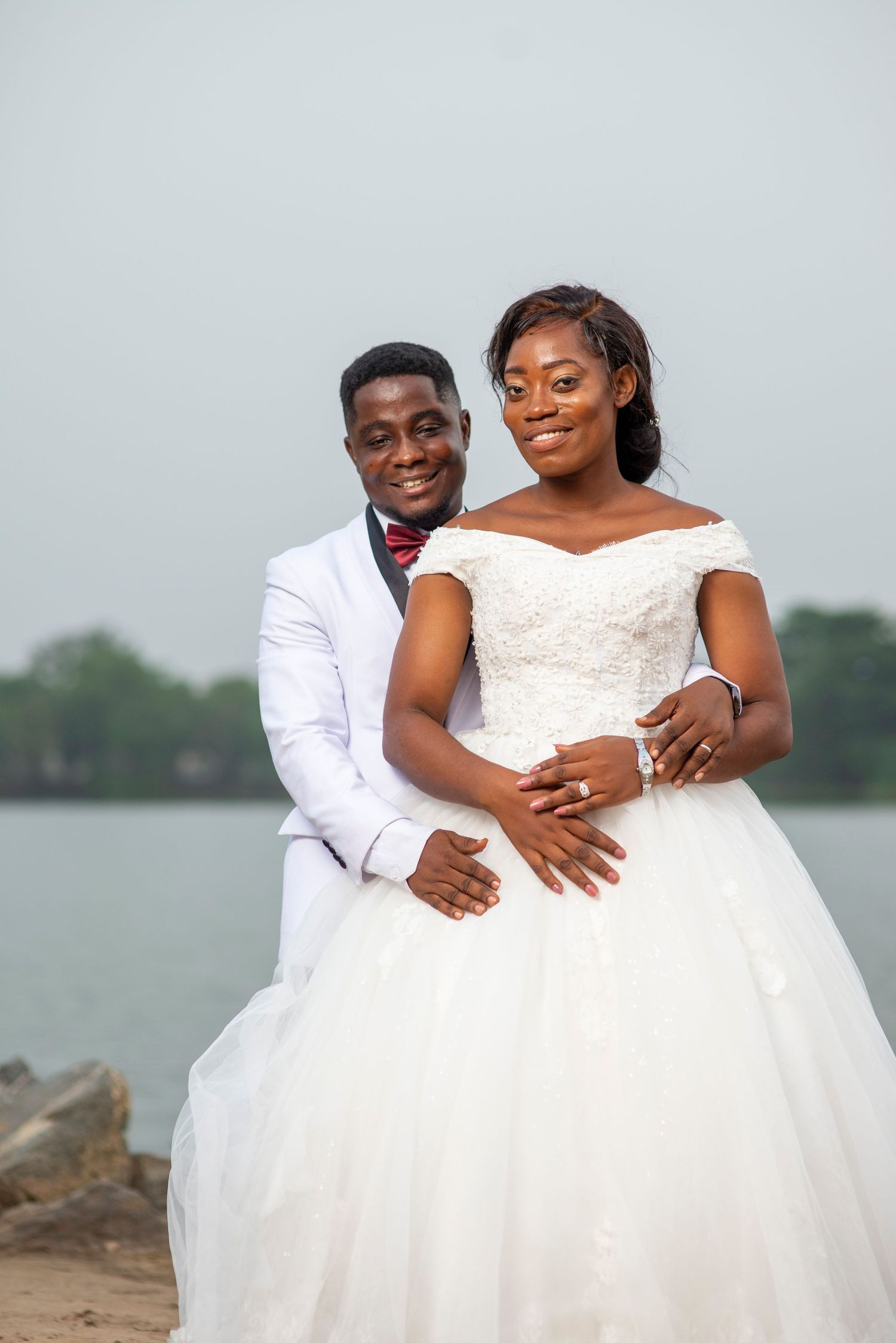 Smiling bride and groom in white wedding attire posing together near scenic waterfront.