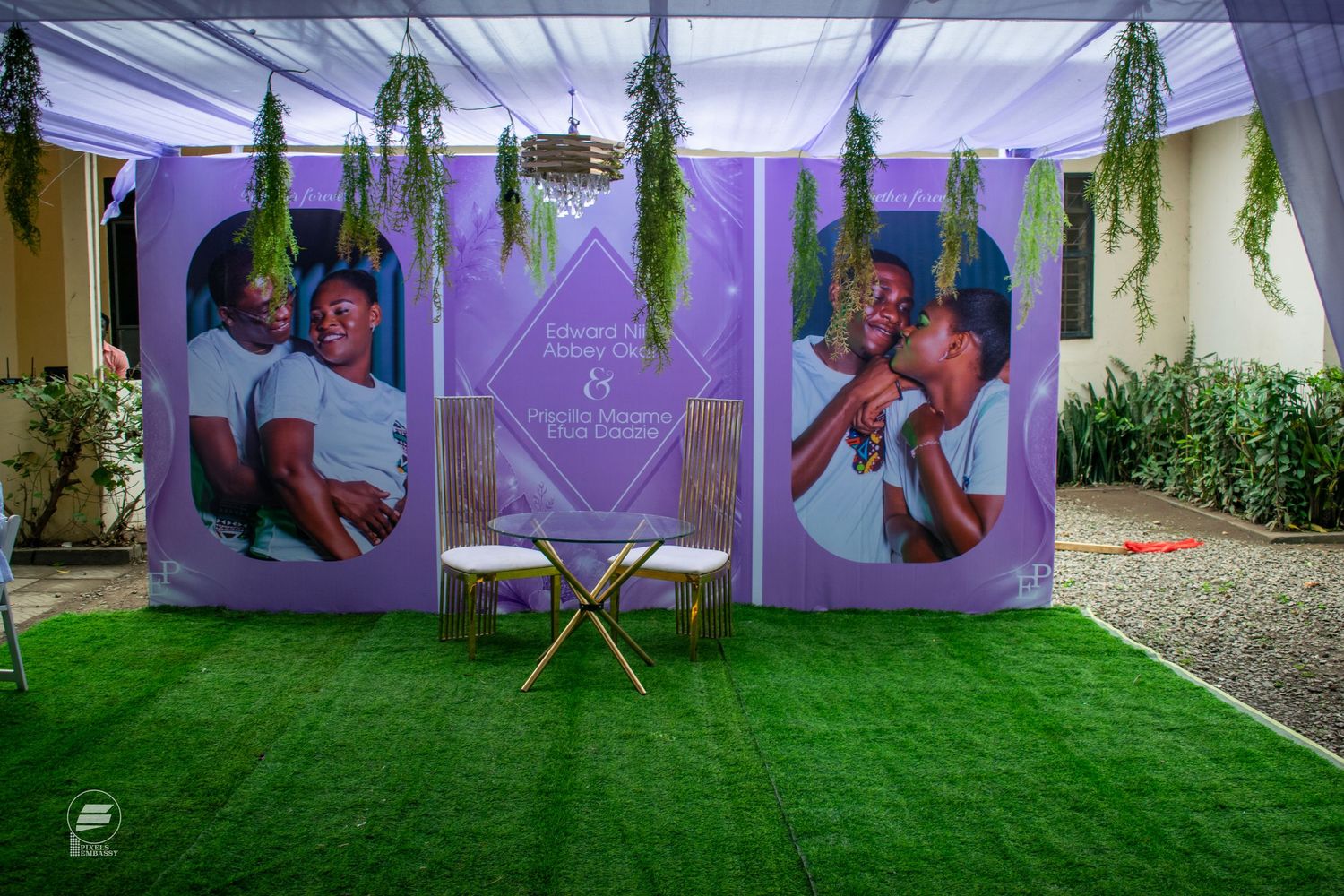 Outdoor wedding backdrop with purple panels featuring couple photos, hanging greenery, and a small gold table on artificial grass.