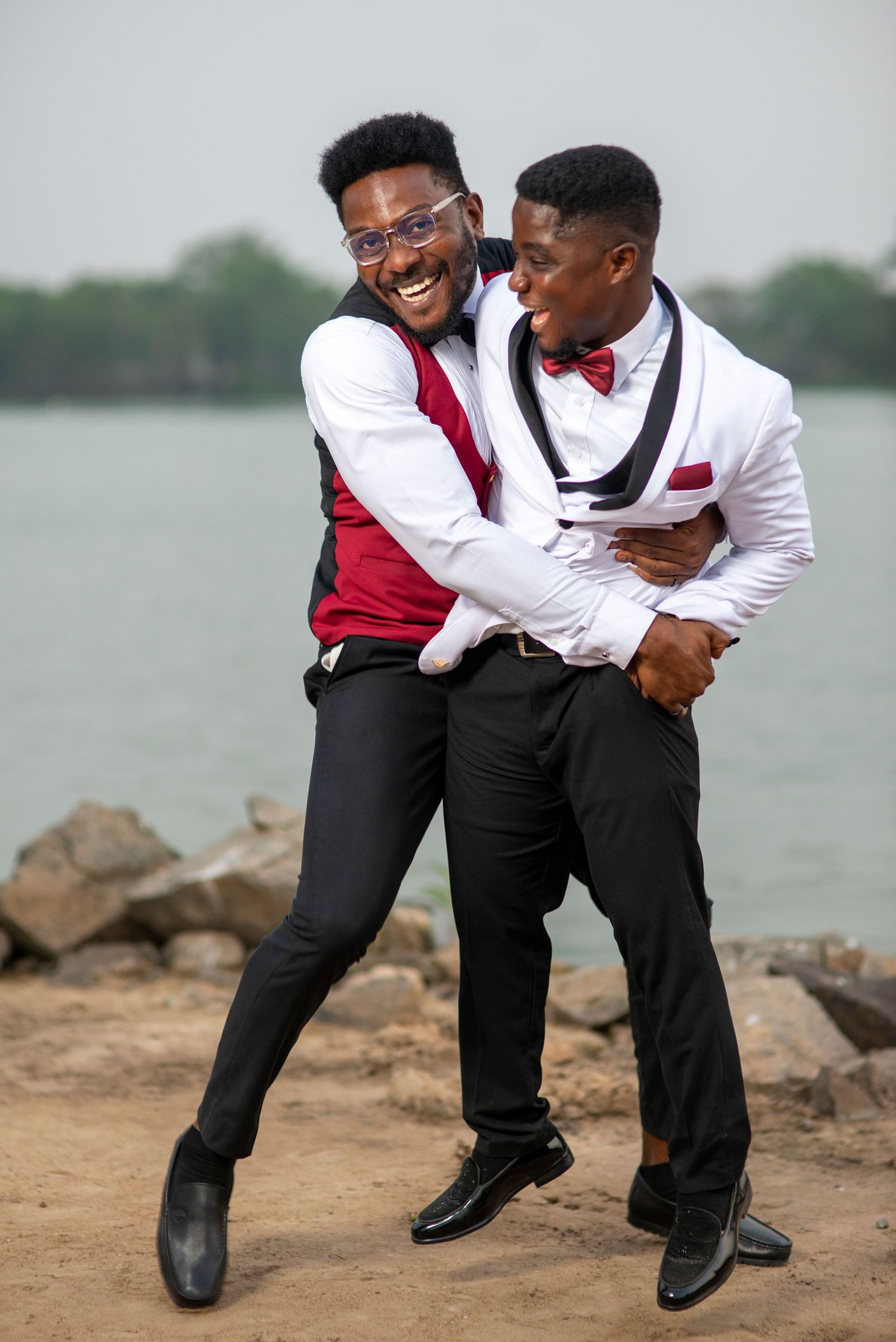 Two well-dressed young men in formal attire laughing and embracing near rocky waterfront.