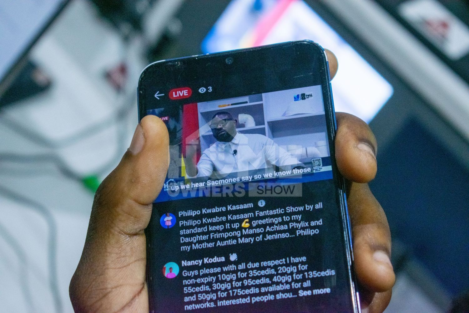 Hands hold a smartphone showing a live Facebook stream of a TV interview with viewer comments visible.