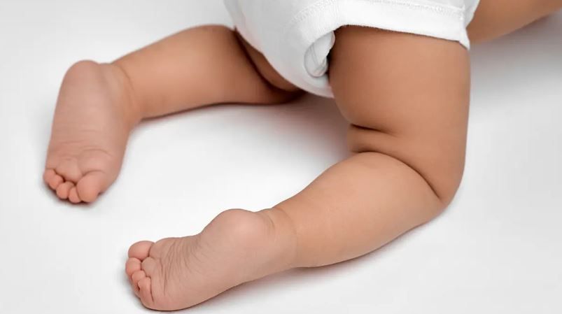 Close-up of chubby baby legs and feet resting on a white surface, wearing a white diaper.