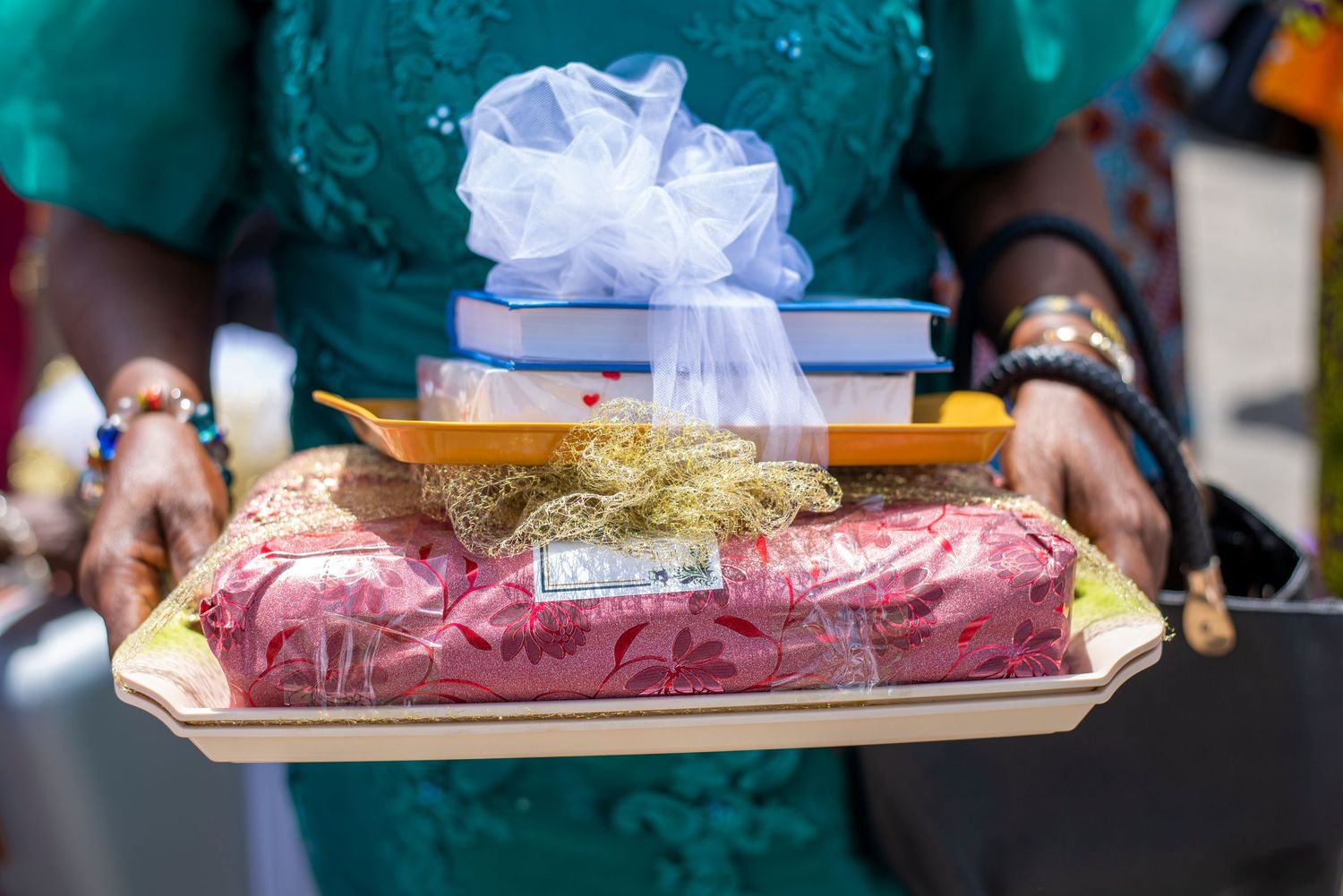 Person in teal outfit holding a decorative tray with wrapped gifts, books, and a white bow at a celebration.