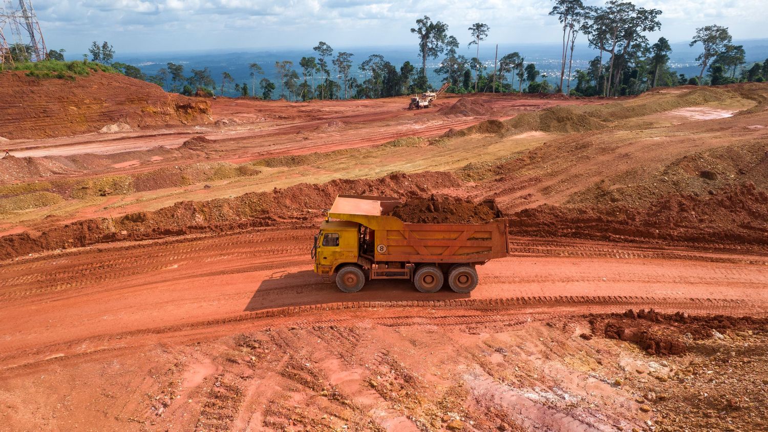 Yellow dump truck hauling red soil on dirt road at open-pit mining site.