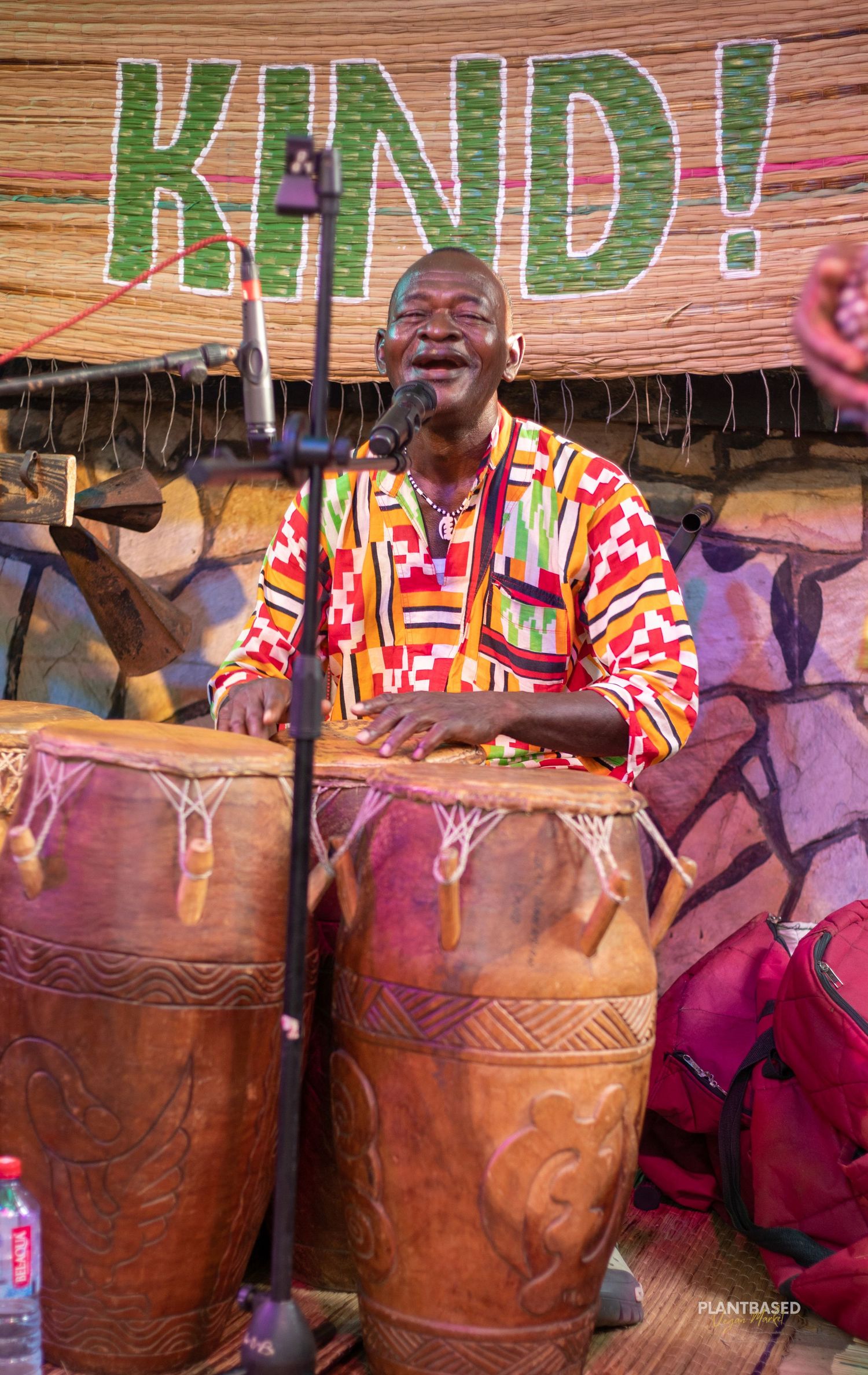 Man in colorful kente shirt playing traditional African drums on stage with KIND sign backdrop.