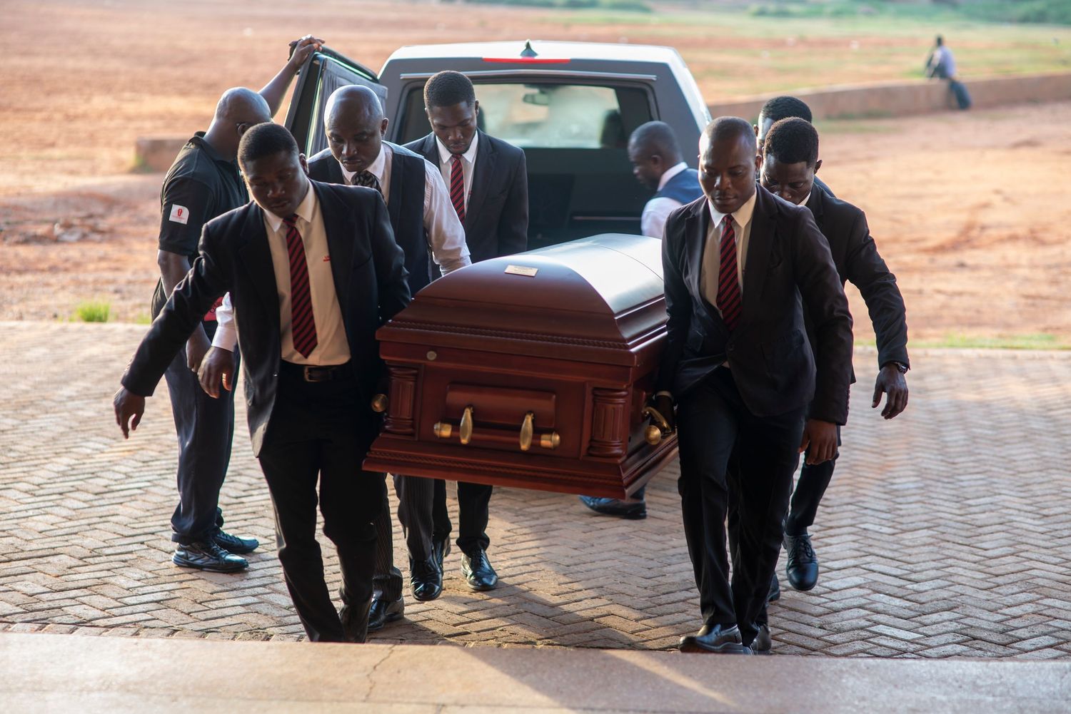 Pallbearers in formal attire carry a wooden casket from a vehicle during an outdoor funeral ceremony.