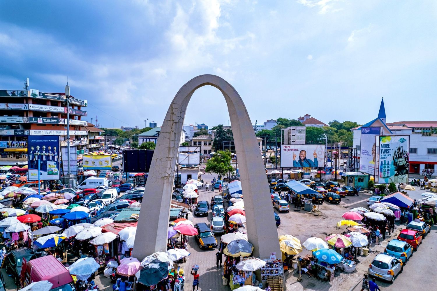 Aerial view of a busy African market with colorful umbrellas surrounding a large concrete arch monument.