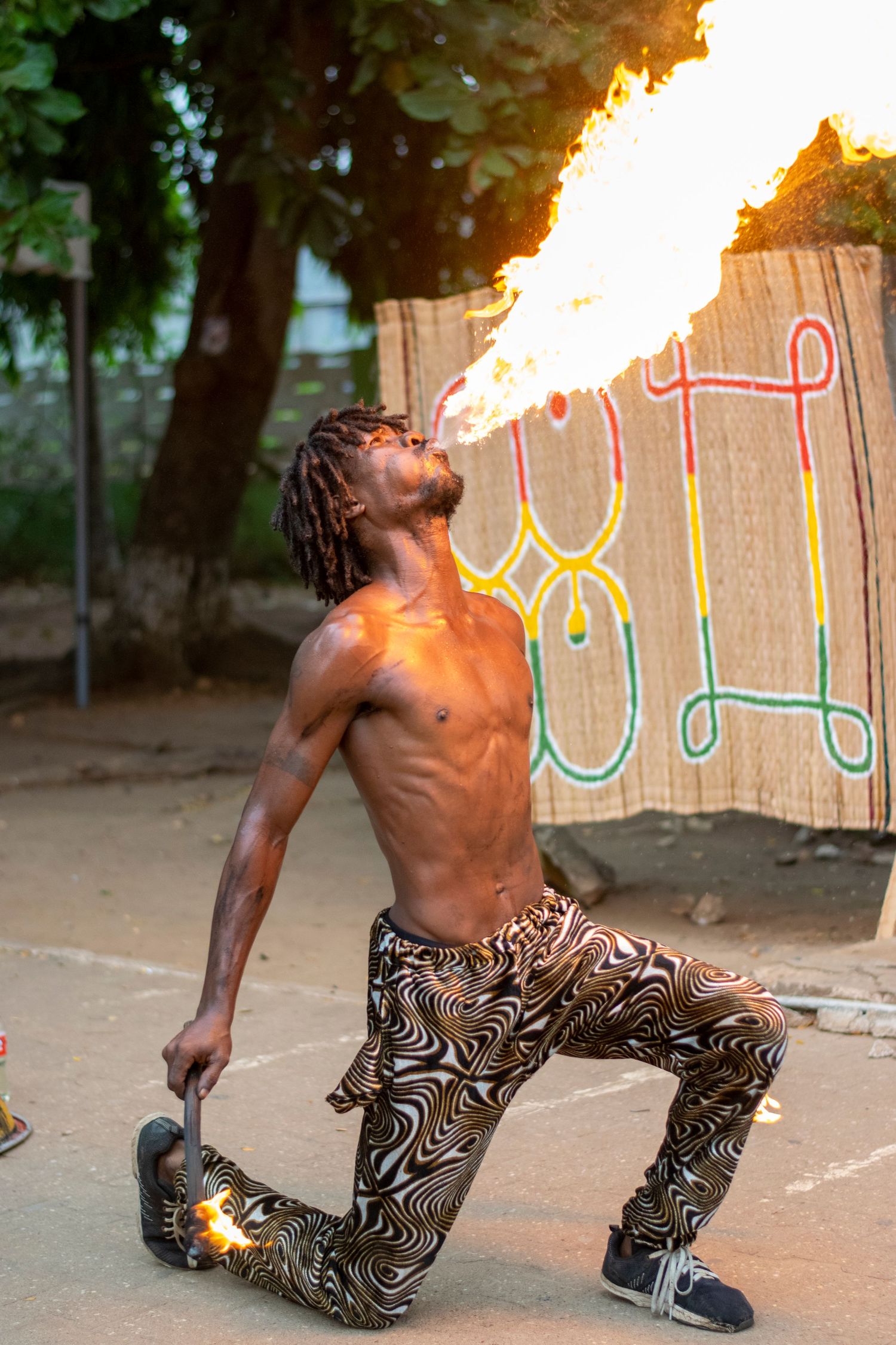 Shirtless African fire breather exhaling a large flame at an outdoor cultural performance.
