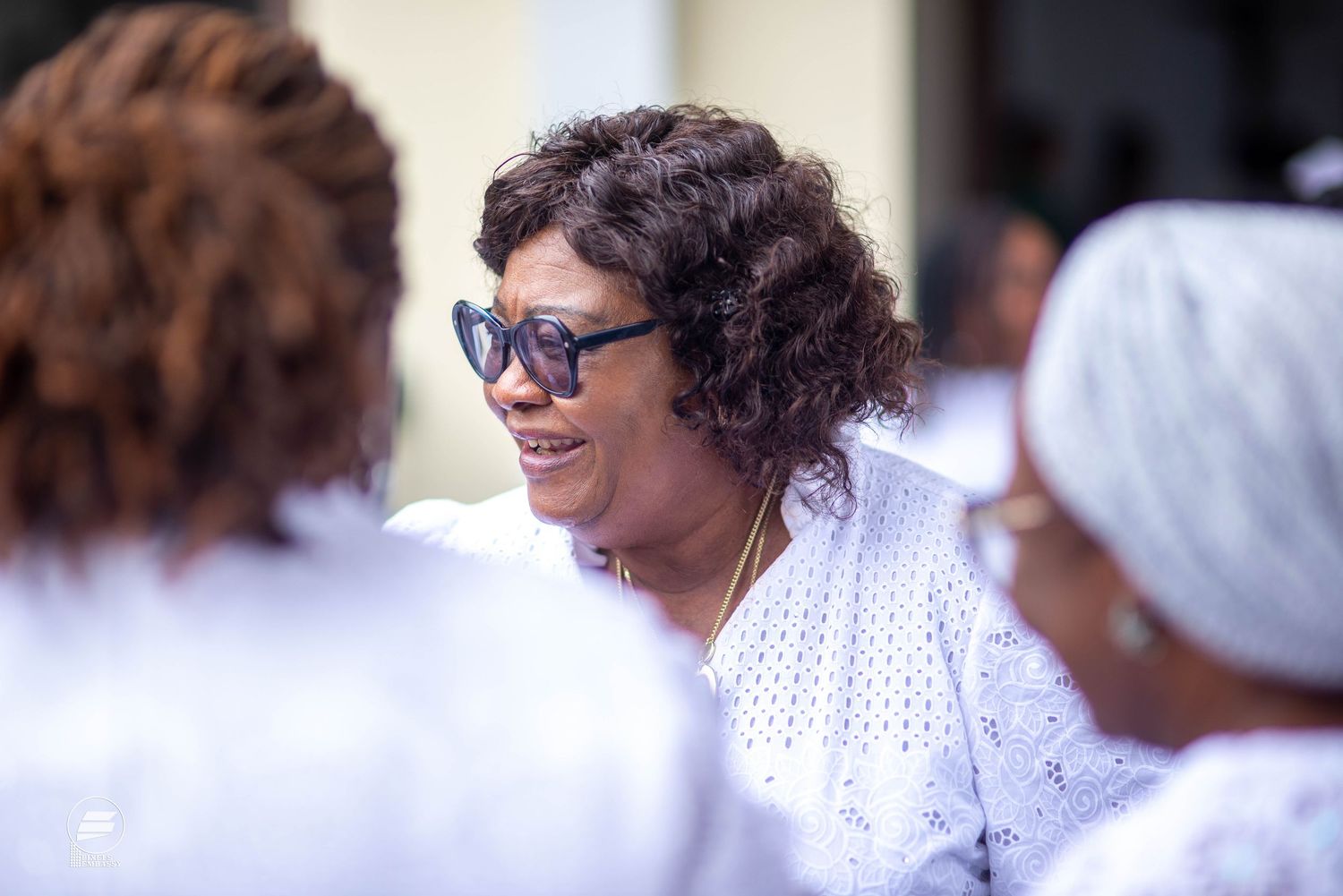 A smiling African woman wearing stylish sunglasses and white lace outfit at an outdoor event.