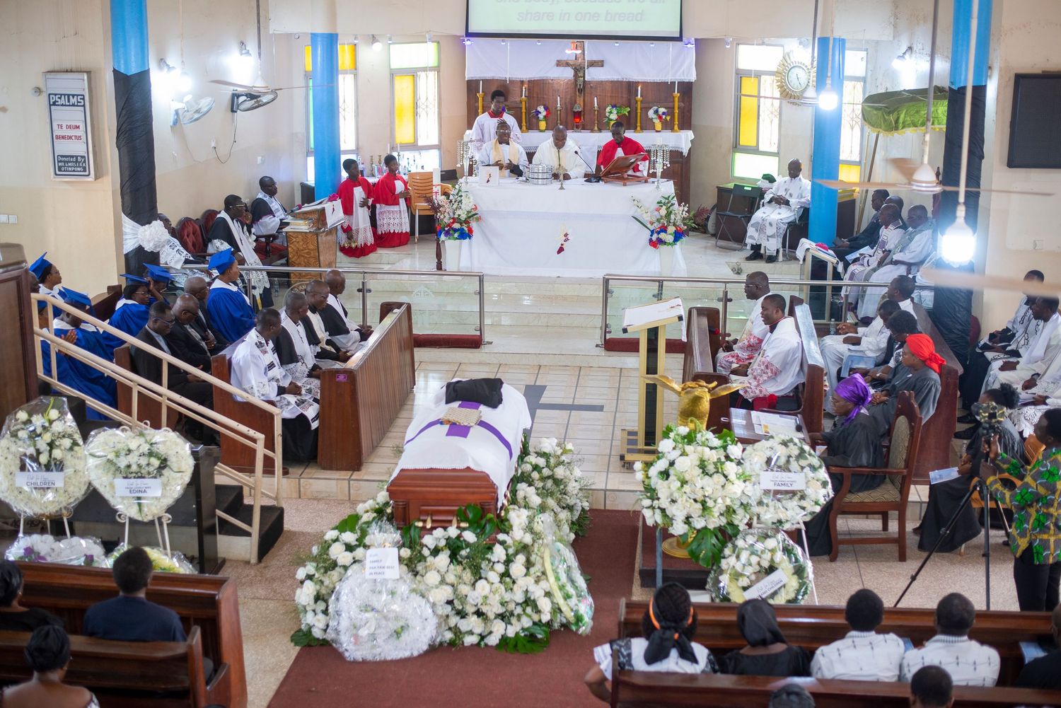 Catholic funeral mass with white flower-adorned casket, mourners in pews, and clergy at the altar in a decorated church.