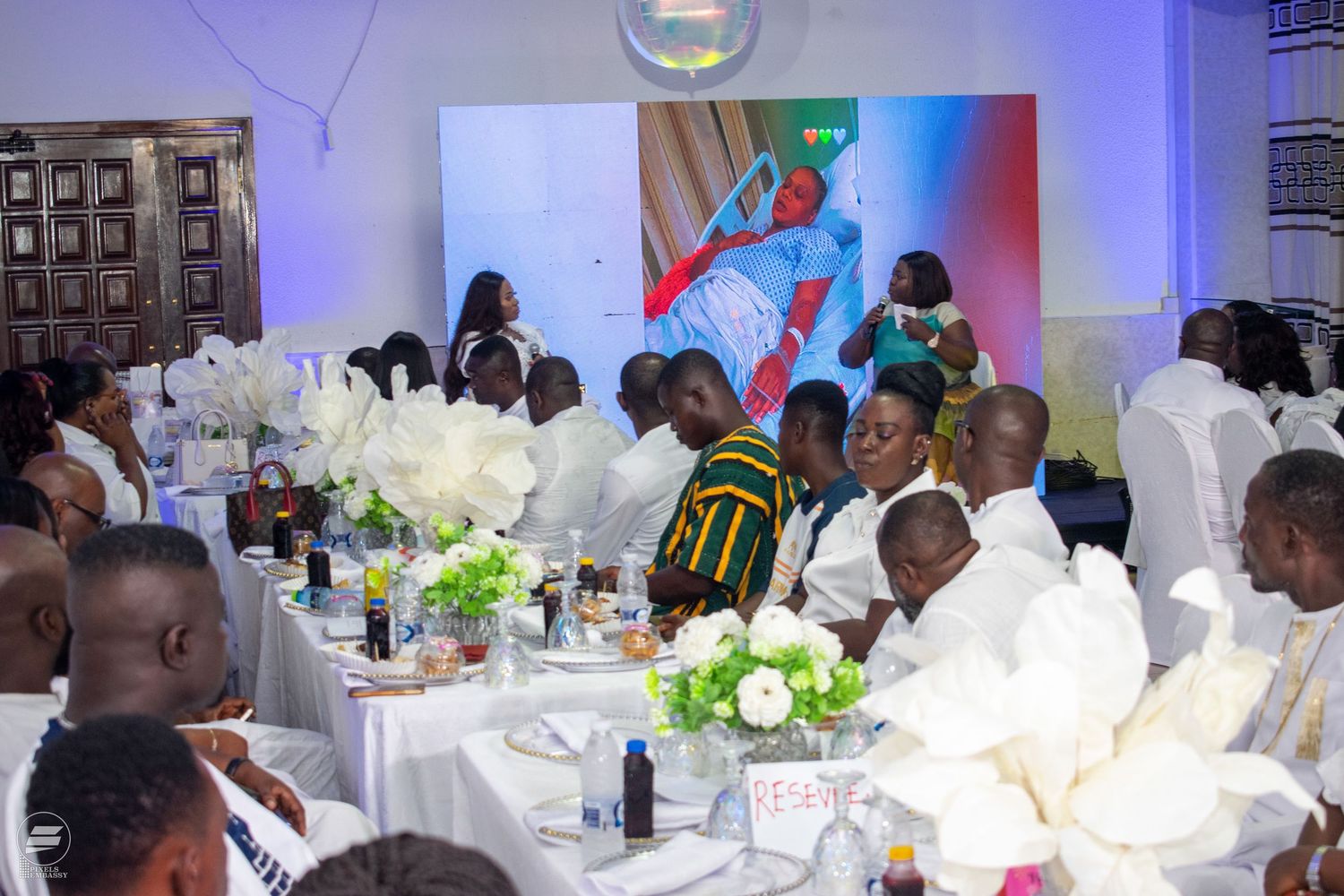 Guests in white attire seated at decorated tables during a formal banquet event with floral centerpieces.