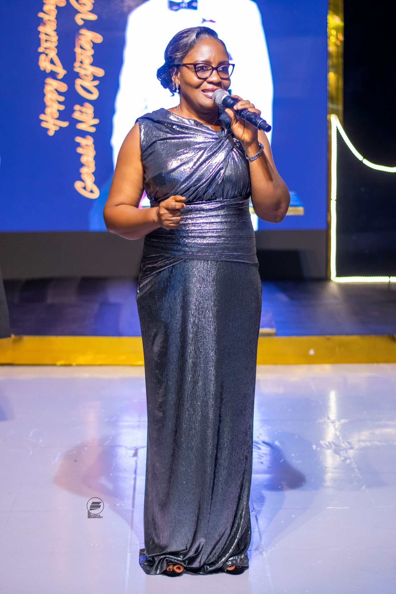Woman in a silver gown speaking into a microphone on a stage at a birthday celebration event.
