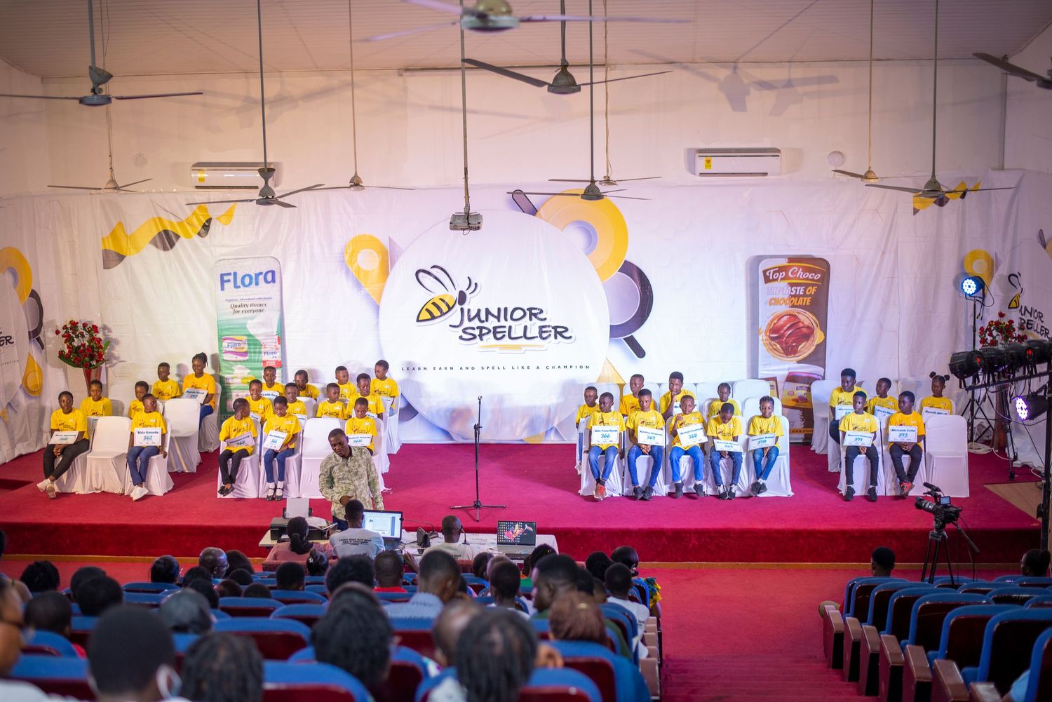 Children in yellow shirts perform on stage at a Junior Speller spelling bee competition event.