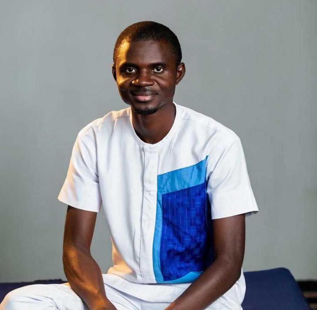 A young African man in a white and blue traditional shirt sitting and smiling confidently.