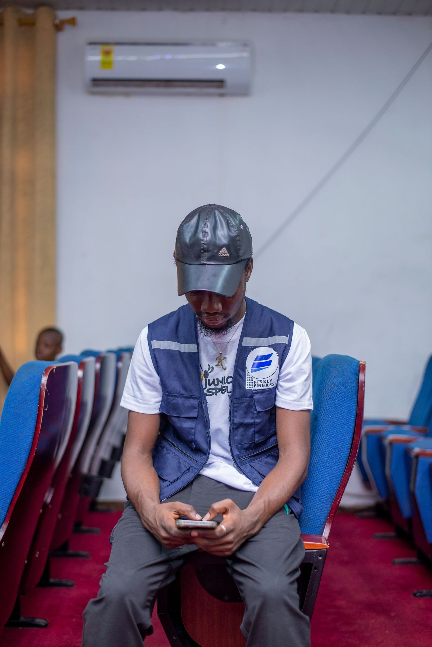 Man in blue vest and cap sitting in blue auditorium chairs looking at his smartphone.