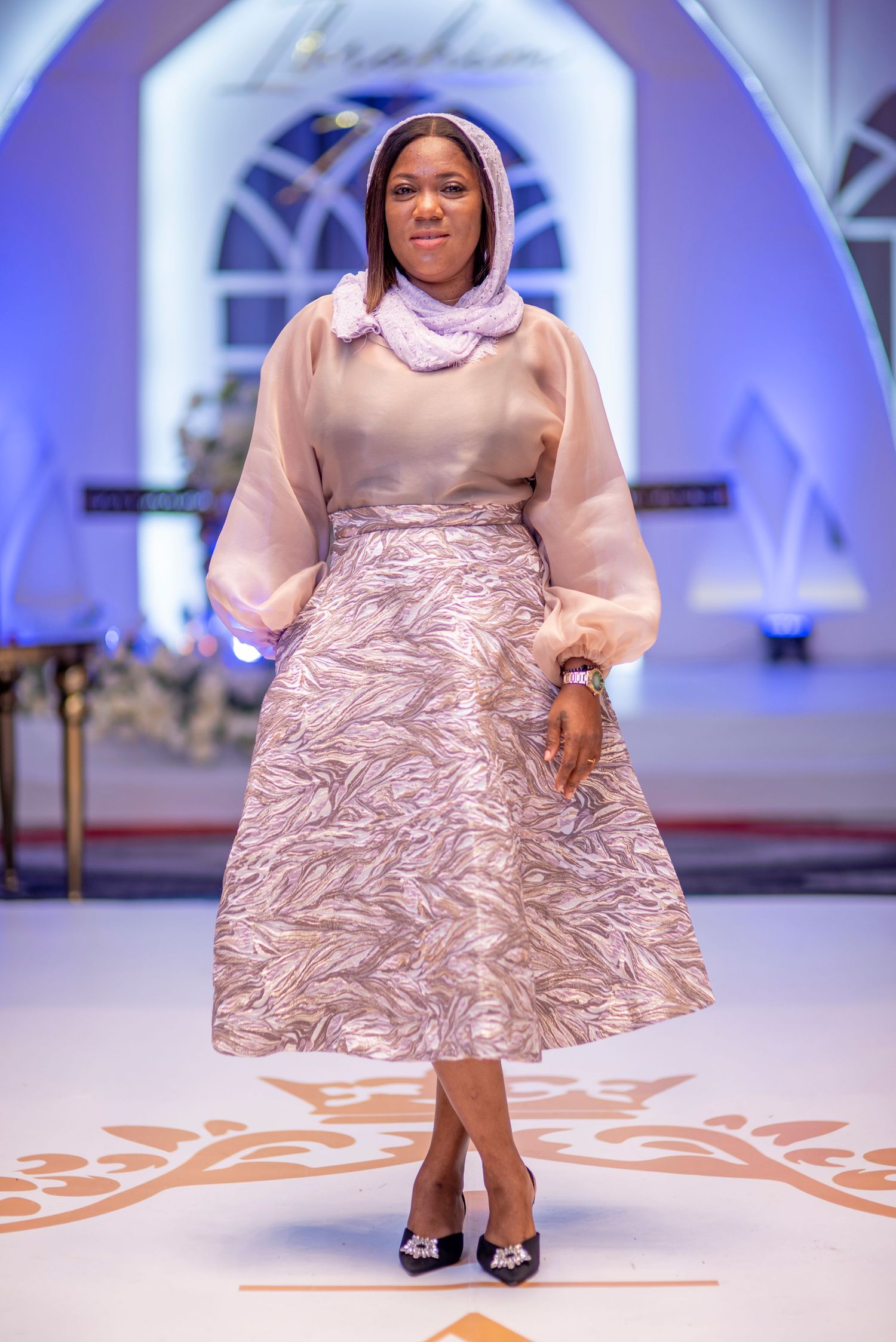 Woman modeling elegant beige midi skirt and sheer blouse on fashion runway.