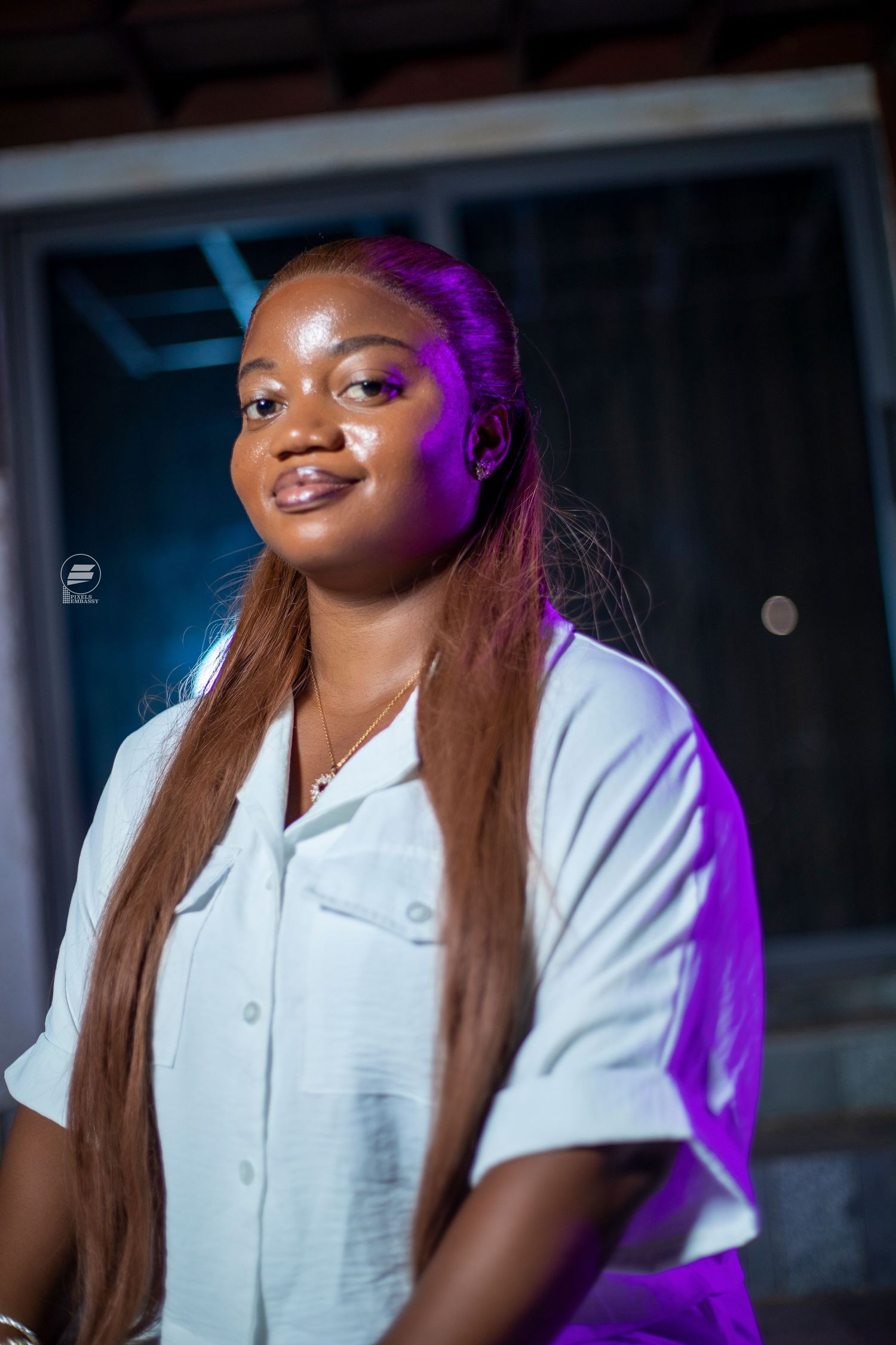 Young woman with long auburn hair in white shirt posing under purple lighting at night.