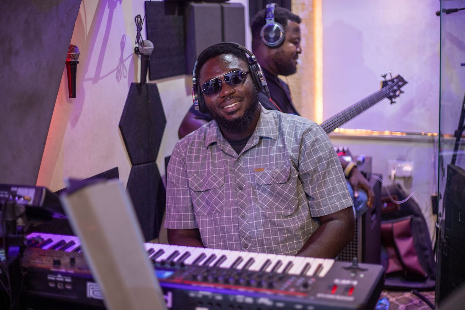 Smiling musician playing keyboard synthesizer in recording studio wearing sunglasses and headphones with guitarist in background.