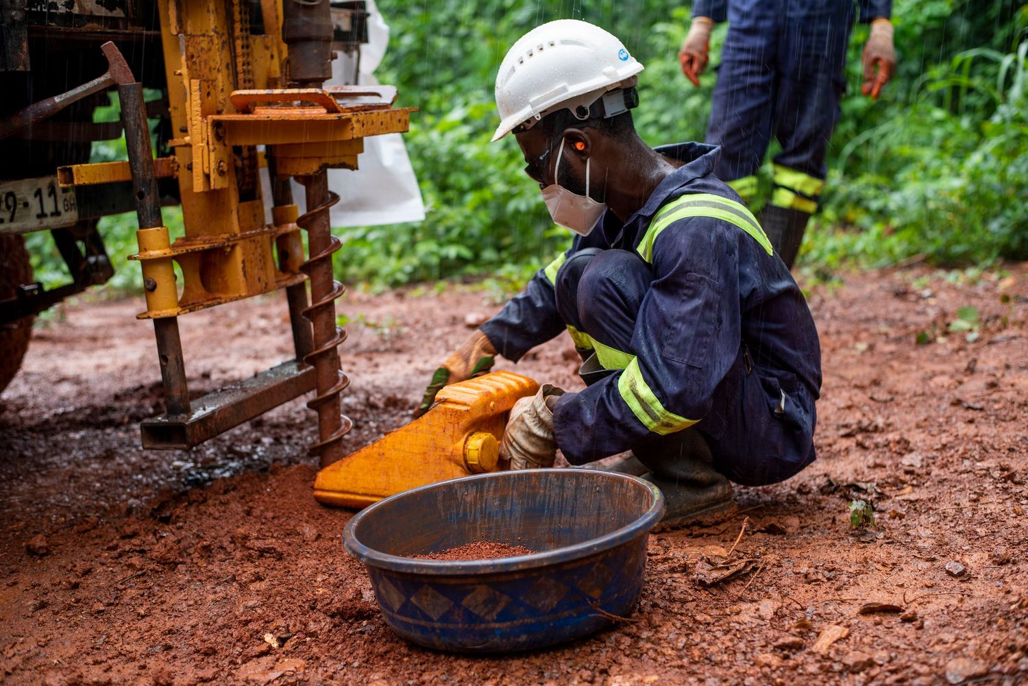Mining worker in safety gear kneeling on red soil collecting drill samples in bowl.