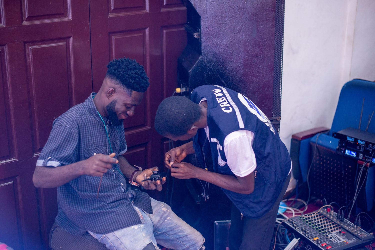 Two men looking at smartphones together near audio equipment by a wooden door.